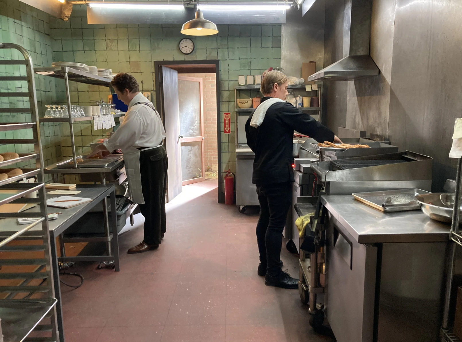 Two cooks working in a commercial kitchen, one chopping and the other grilling food, with shelves of dishes and racks of bread visible.