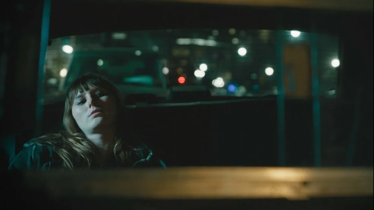 A woman sitting in the backseat of a vehicle at night, looking tired or upset, with city lights blurred in the background.