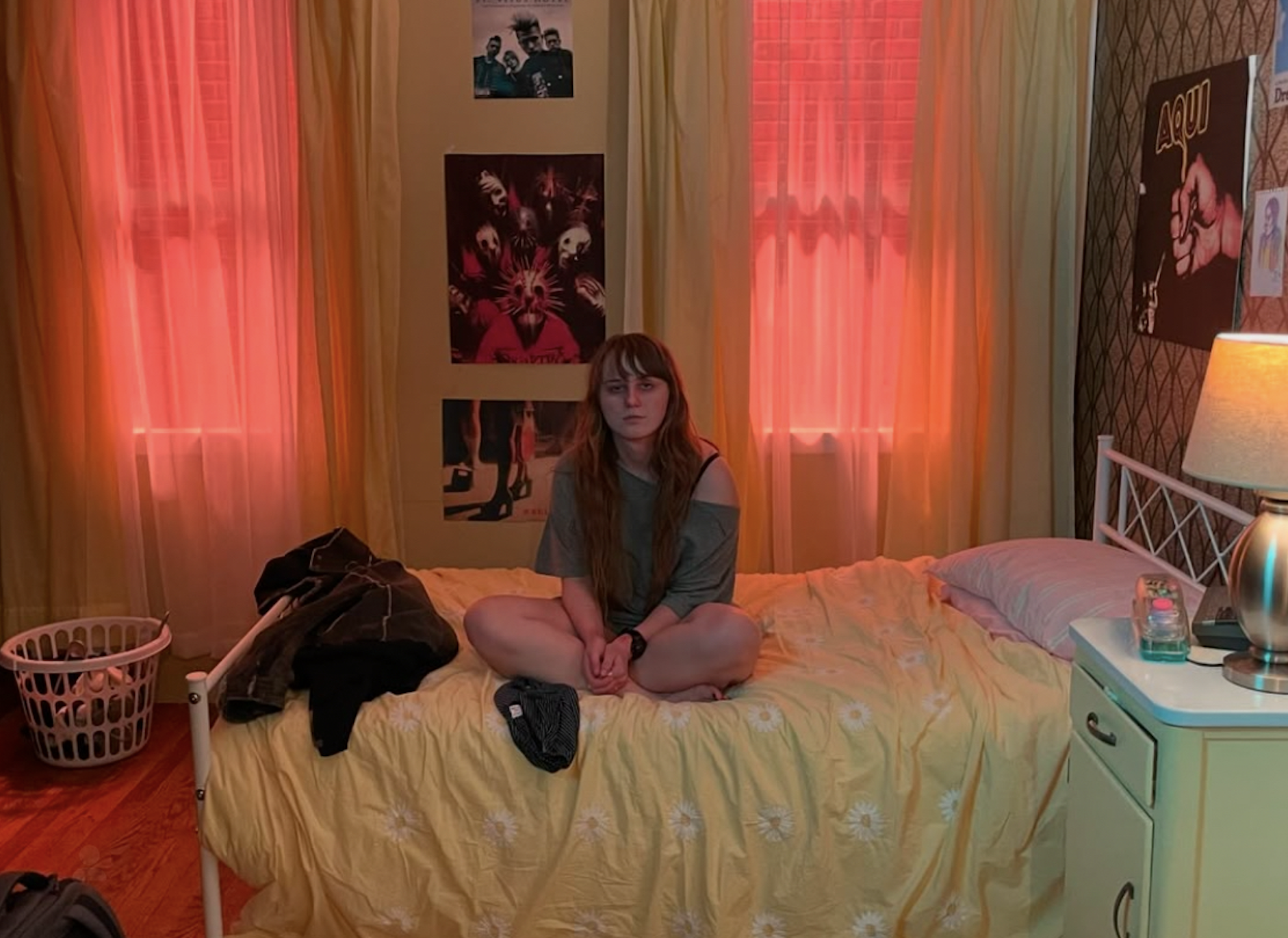 Young woman sitting on a bed in a dimly lit bedroom with orange curtains, posters on the wall, a bedside lamp, and a laundry basket.
