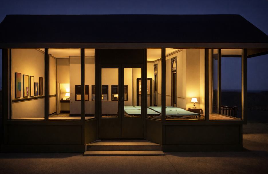 Inside a well-lit modern living room at night, with large glass windows showcasing a dark landscape, featuring a couch, art on the wall, and lamps on side tables.