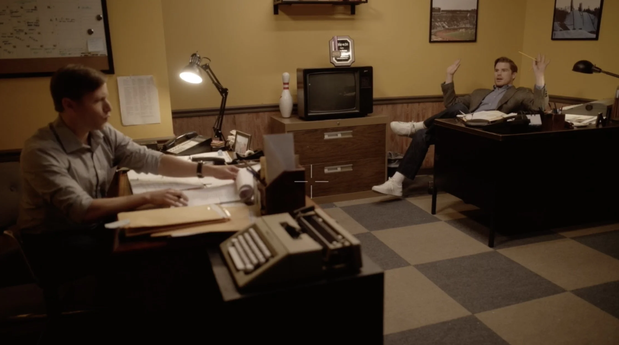 An office scene with two men: one sitting at a desk with papers and a typewriter, the other sitting relaxed in a chair with legs up on the desk, gesturing with hands. The background includes a TV, a bowling pin, and framed pictures on the yellow walls.