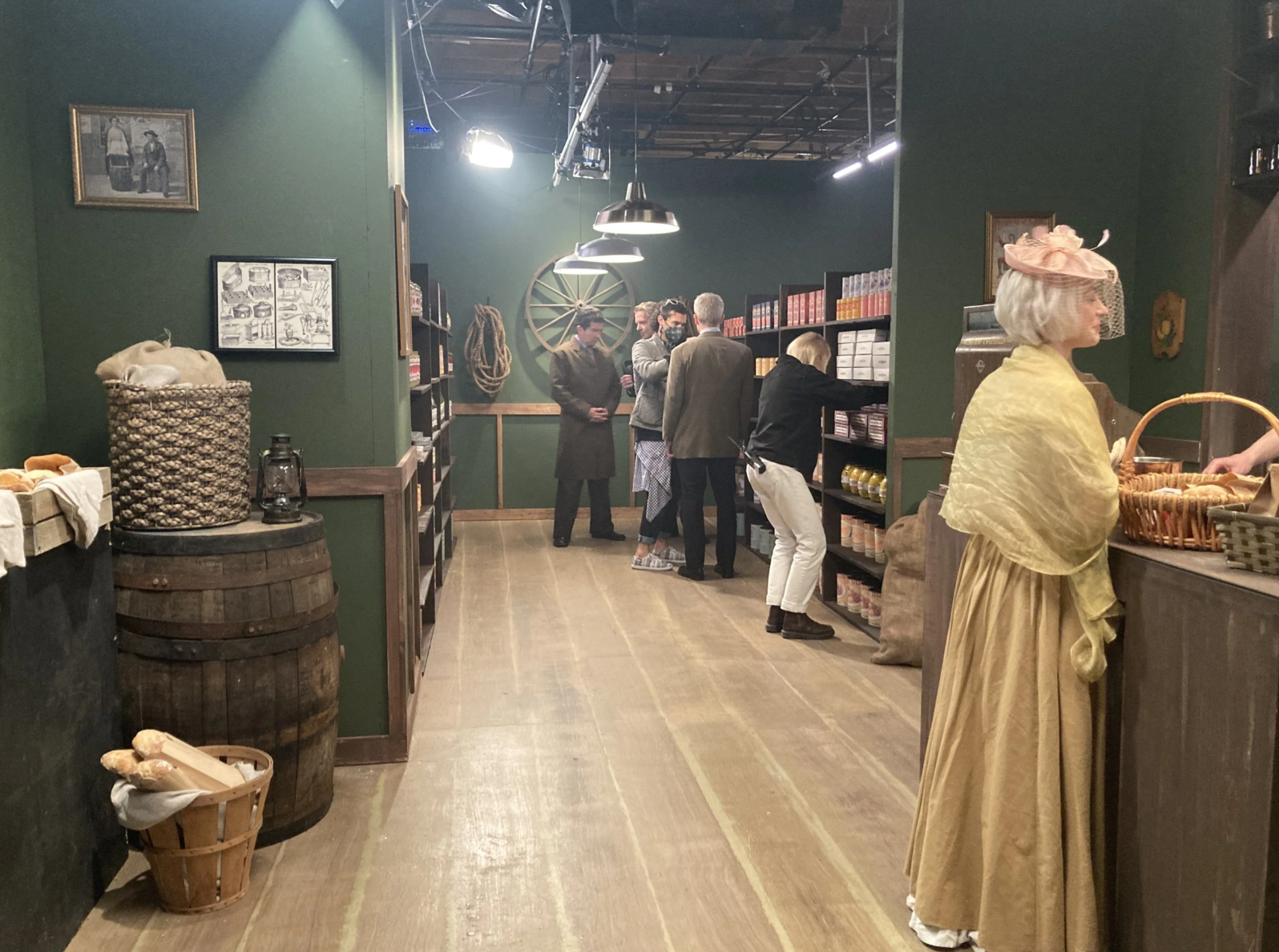 People shopping in a store with wooden shelves and green walls, vintage decor, and a woman in historical costume in the foreground.