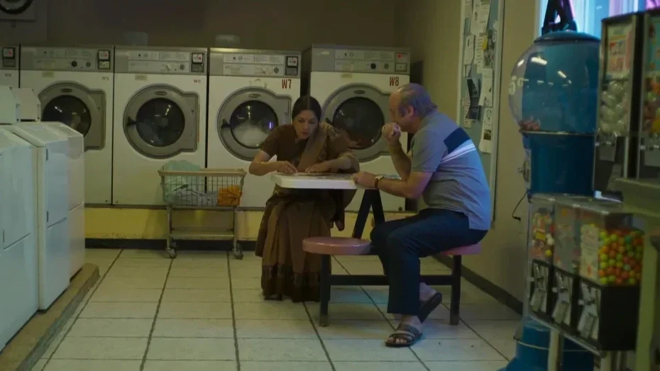 A woman and a man sitting at a small table in a laundromat, with washing machines in the background and a gumball machine on the right side.
