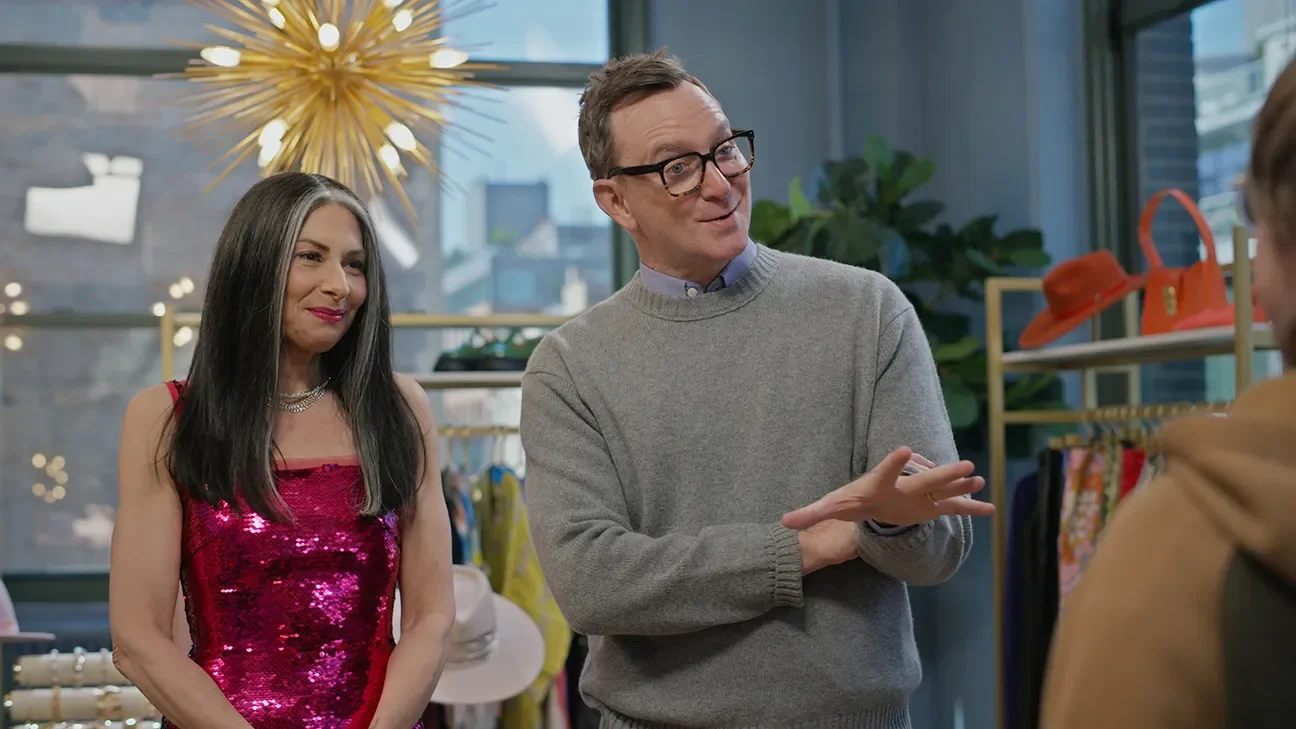 A man and a woman are talking inside a store, with the woman wearing a shiny pink dress and the man wearing glasses and a gray sweater. There is a large decorative light fixture above them and store shelves displaying clothing and accessories in the background.
