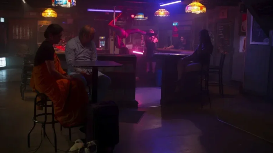 A dimly lit bar with neon and colored lighting, featuring a pole dancer performing an aerial acrobatic routine, and several patrons seated at the bar or tables, including two women in conversation.