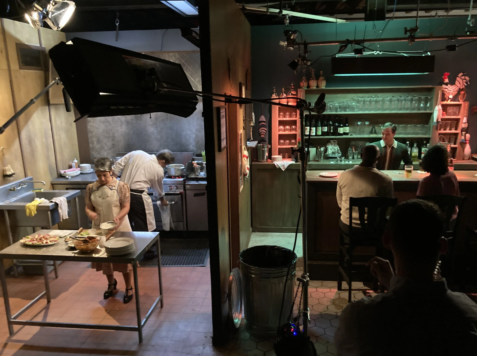 A restaurant kitchen on the left with chefs preparing food, and a bar area on the right with patrons sitting and a bartender behind the bar, with dim lighting and holiday decorations.