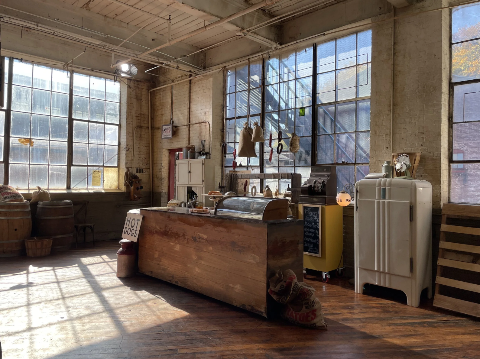 Interior of a rustic cafe or market with large industrial windows, wooden floors, a counter with a display case, and various hanging bags and tools.