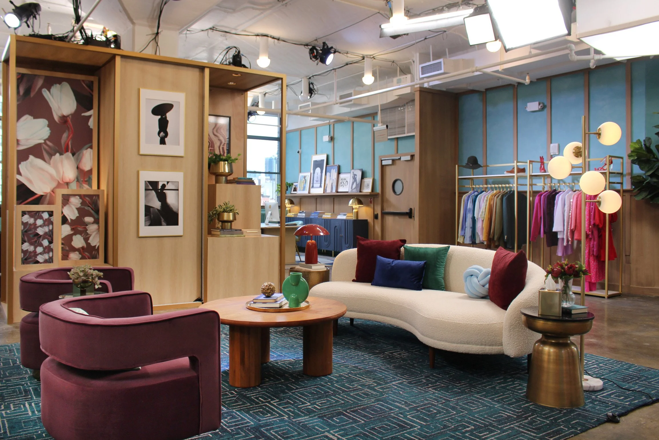 Interior of a modern, stylish boutique clothing store with colorful clothes on racks, a cozy seating area with velvet chairs and a sofa, decorative lighting, artwork, and plants.