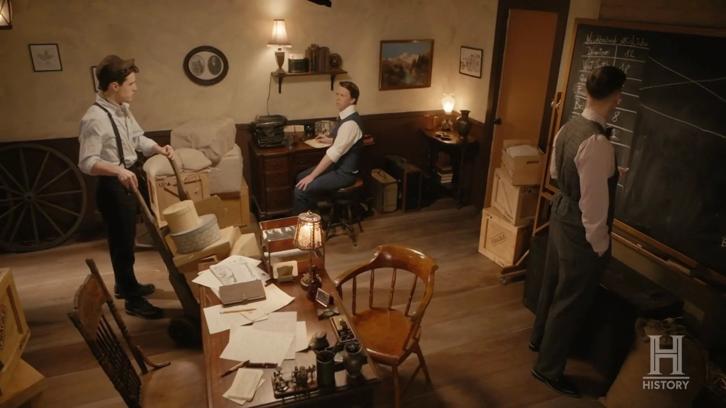 Three men in a vintage-style office or study, one standing with a cart of boxes, one sitting at a desk looking at a computer, and one writing on a blackboard covered with charts or graphs. The room has wooden floors, desk, shelves, and framed pictures on the wall.