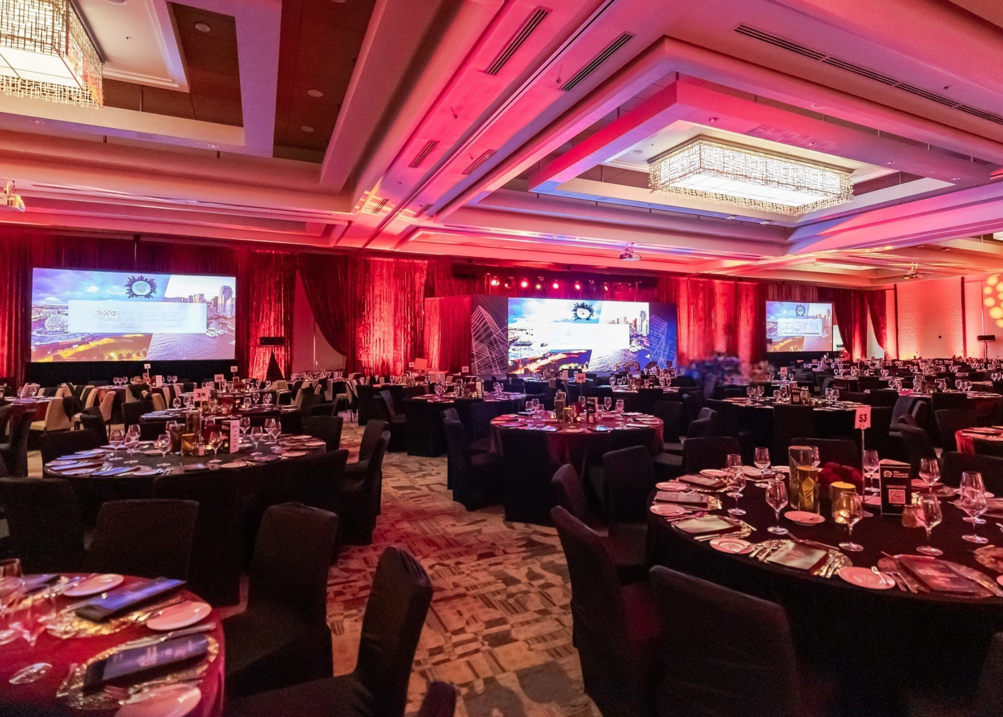 A large banquet hall set up for an event, with round tables covered in red tablecloths, black chairs, and multiple large screens displaying a cityscape with a logo, accented by pink and red lighting.