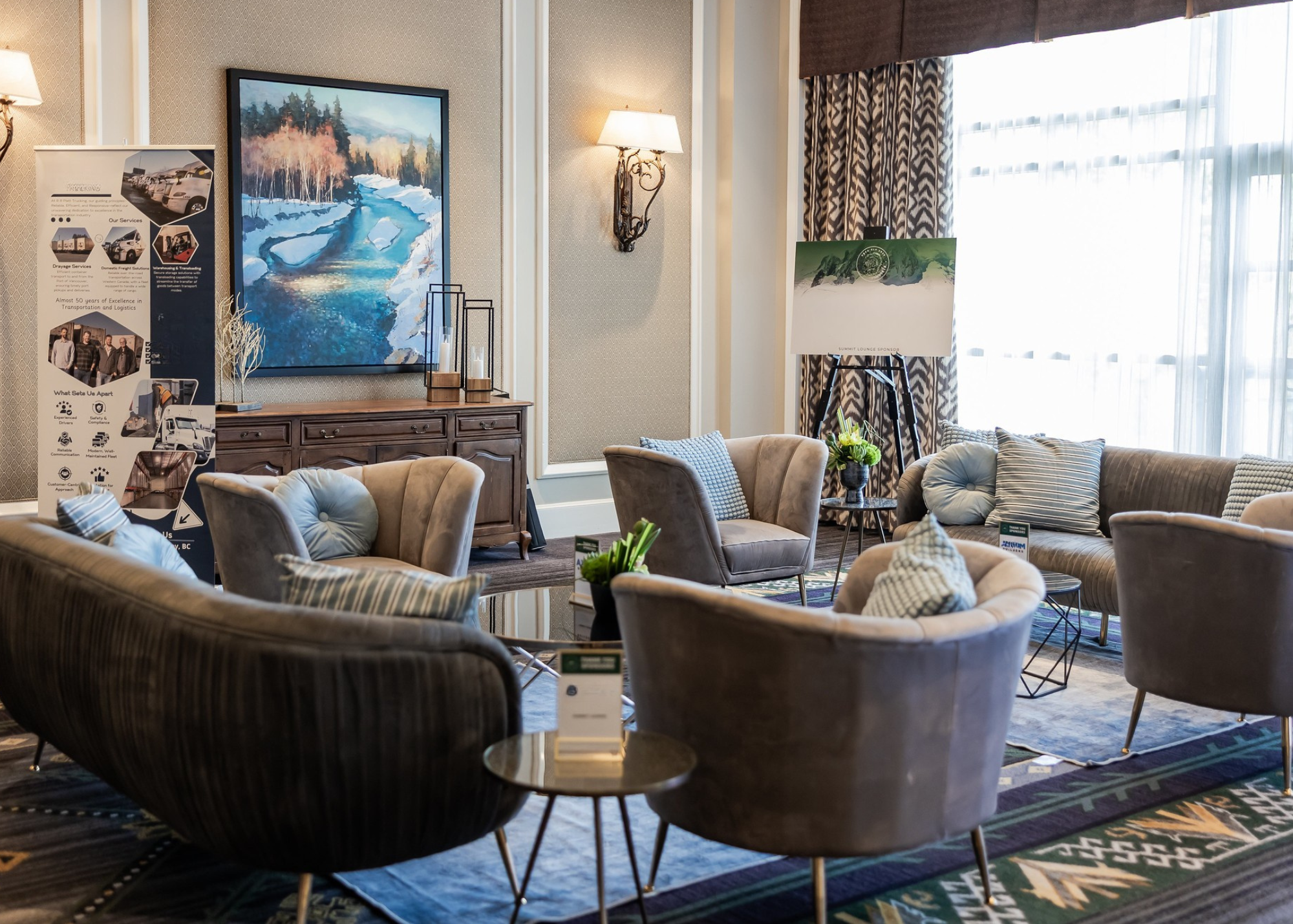 A cozy hotel lobby with beige and gray armchairs and a couch, a wooden sideboard with art and decorative items, a large landscape painting, a standing floor lamp, and a window with curtains.