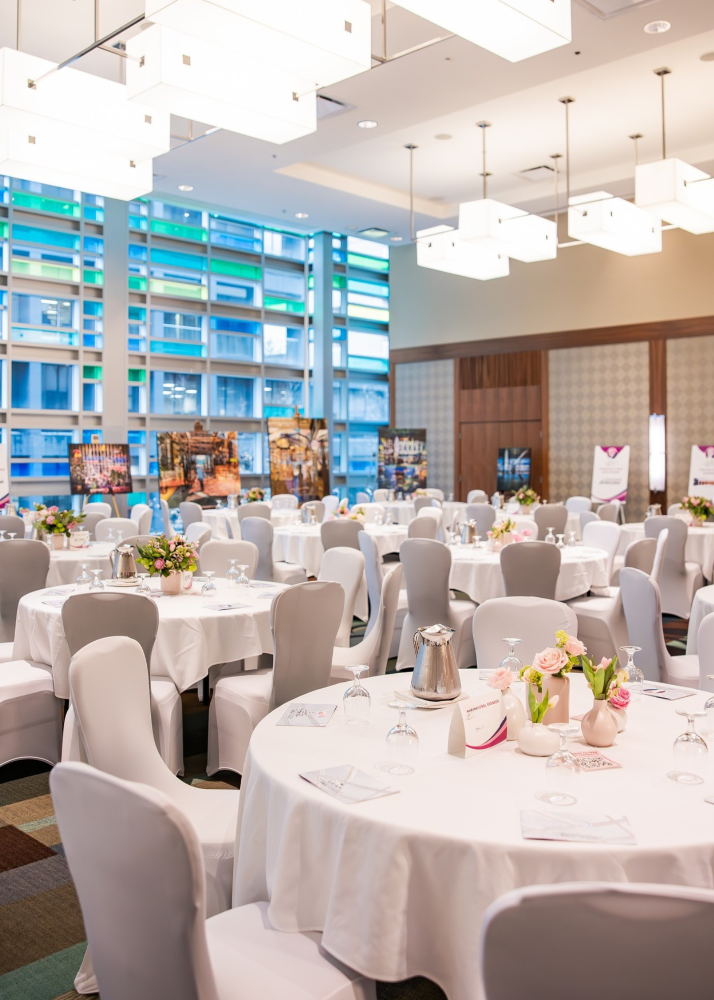 A banquet hall with round tables covered in white tablecloths, decorated with pink and green floral centerpieces, set with glassware and menus. The hall features large colorful window panels and modern white overhead lighting.