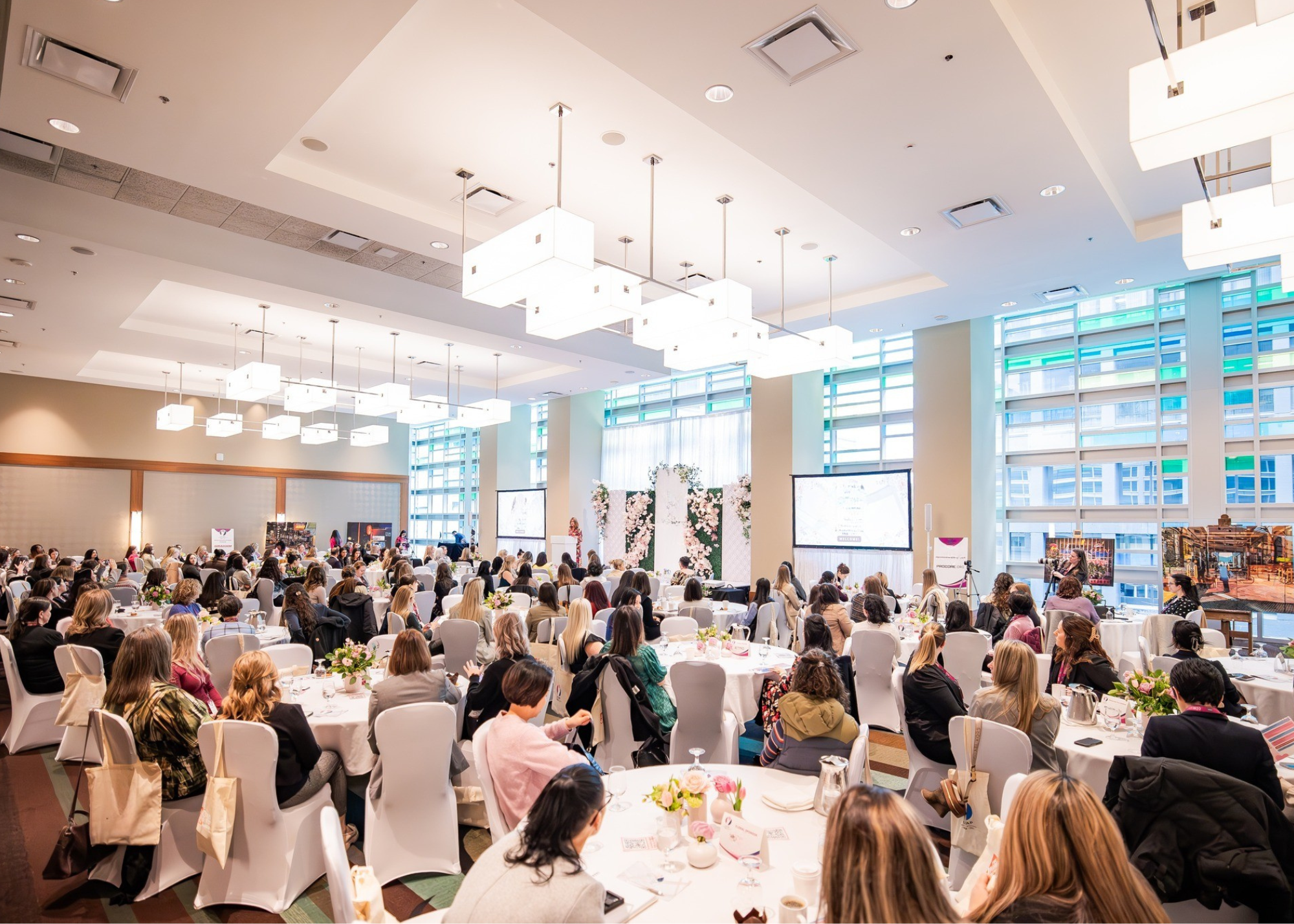 A large conference room filled with rows of round tables decorated with pink and white flower centerpieces. Many women, mostly dressed in business attire, are seated and appear to be listening to a speaker at a podium. The room has high ceilings with