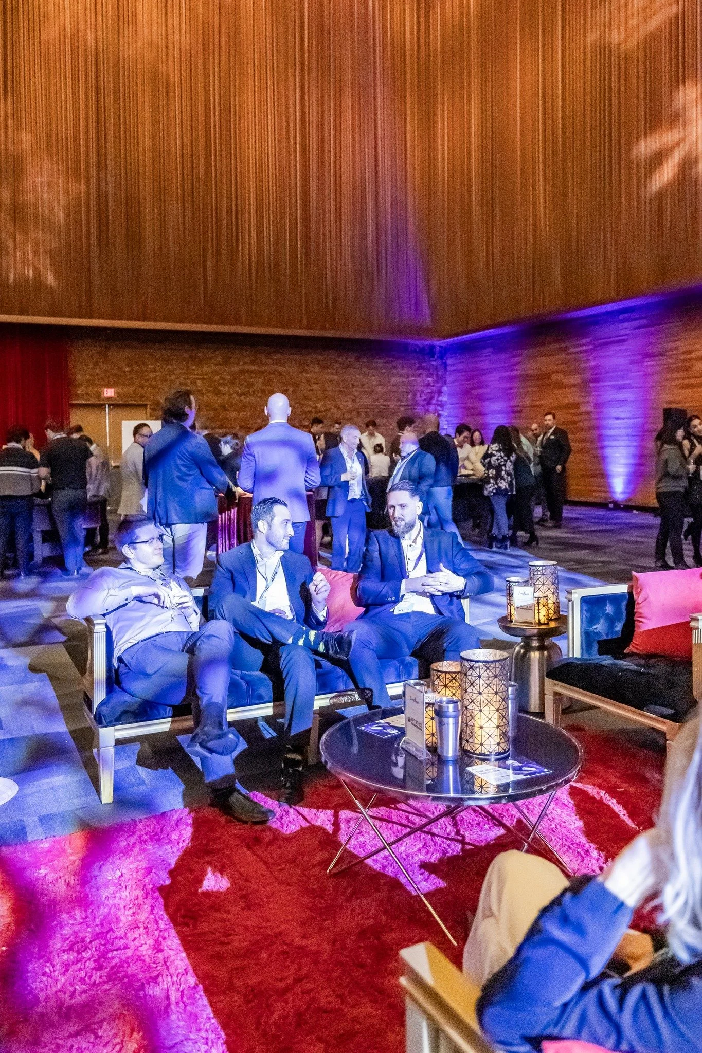 People networking and socializing at a formal event in a modern, wooden, high-ceilinged venue with purple ambient lighting. The foreground shows three men in suits sitting on a sofa.
