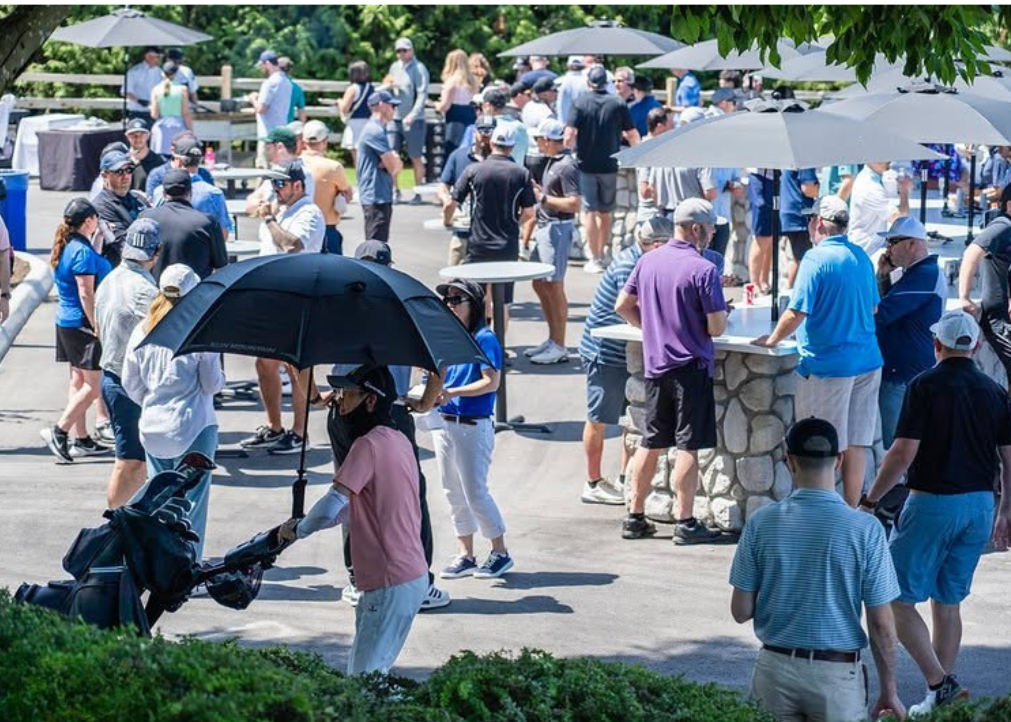 Crowd of people gathered outdoors at a social event on a sunny day, with some under umbrellas for shade. Some individuals are talking at tables, while others are walking around or standing in groups. The setting appears to be a park or outdoor venue 