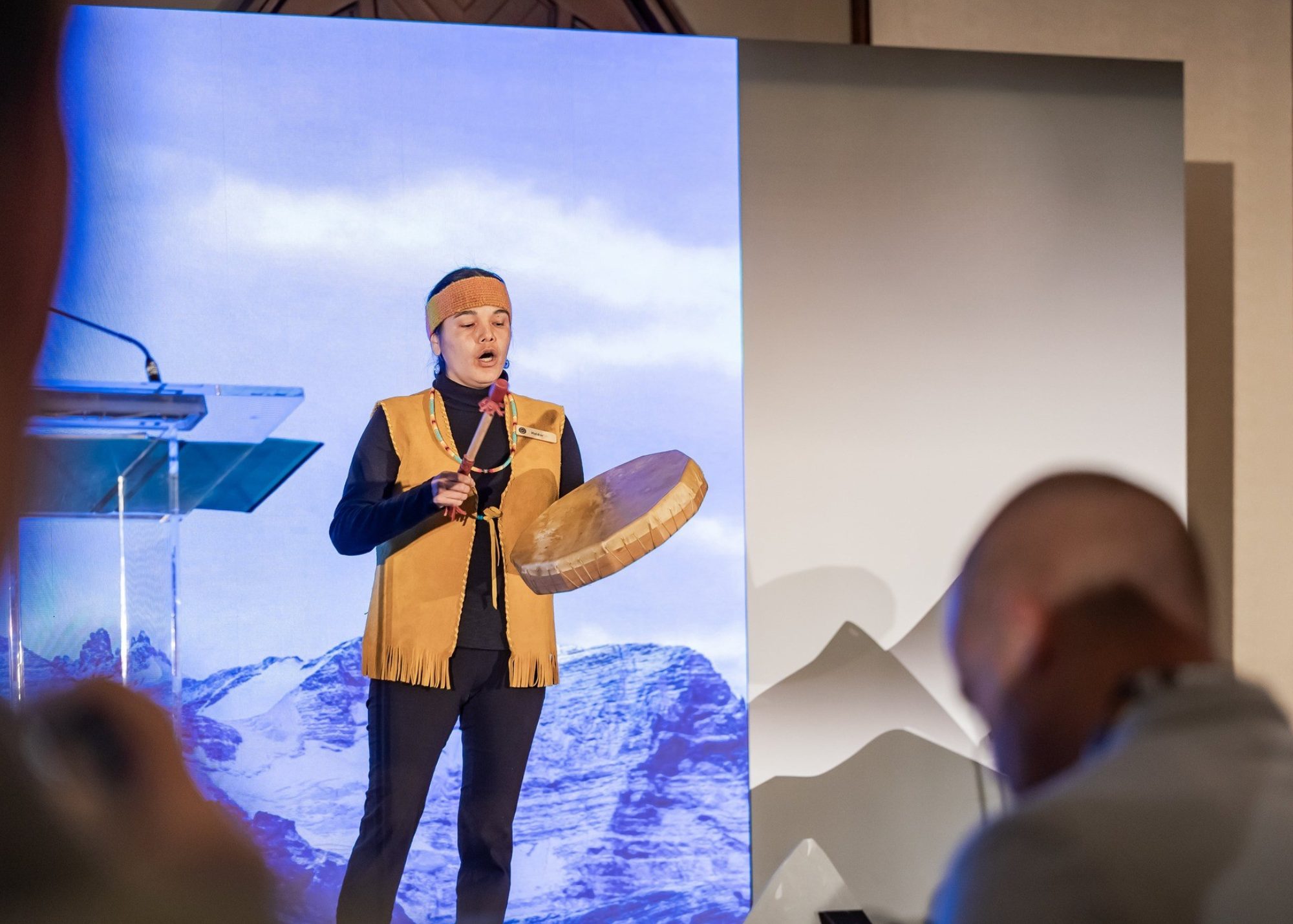 A woman dressed in traditional indigenous clothing, including a fringed vest and beaded necklace, is playing a drum and singing on stage during a conference or performance, with a mountain landscape background on a large screen behind her.