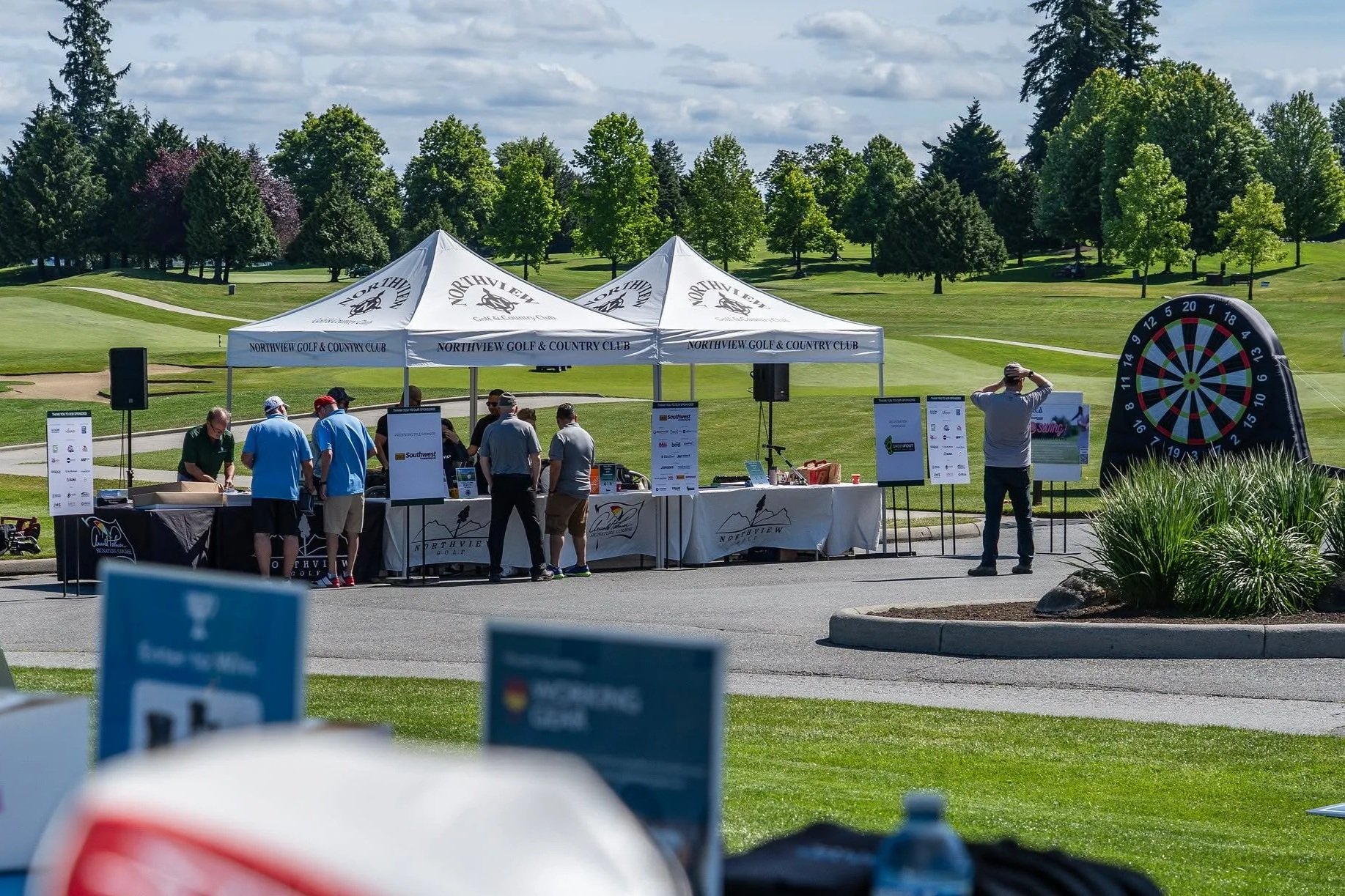 A group of people gather under a white tent at a golf course, with a large inflatable dartboard and a landscaped area with bushes nearby on a sunny day.