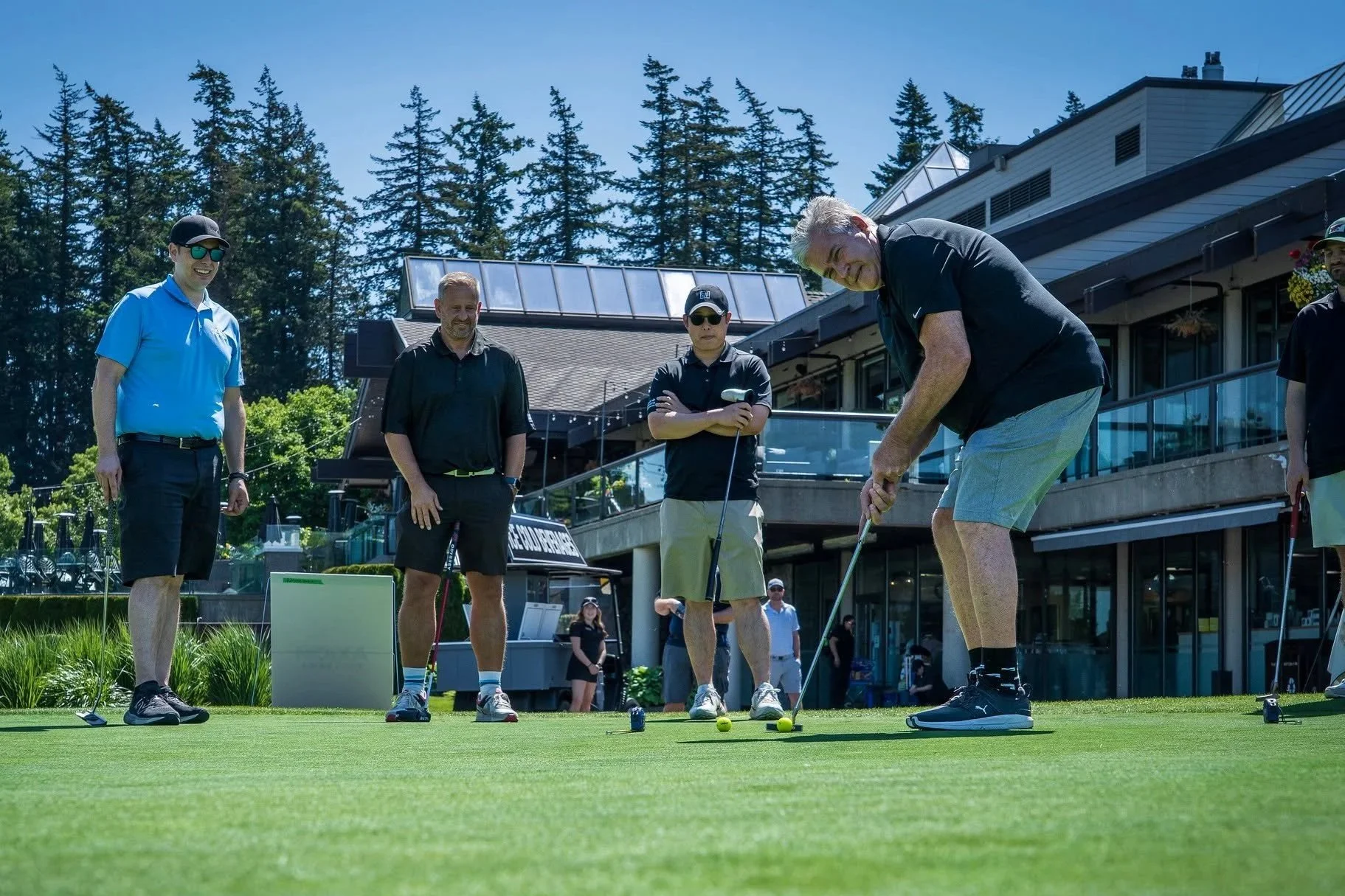 A group of men playing golf on a sunny day, with some standing and watching, on the green near a clubhouse with trees in the background.