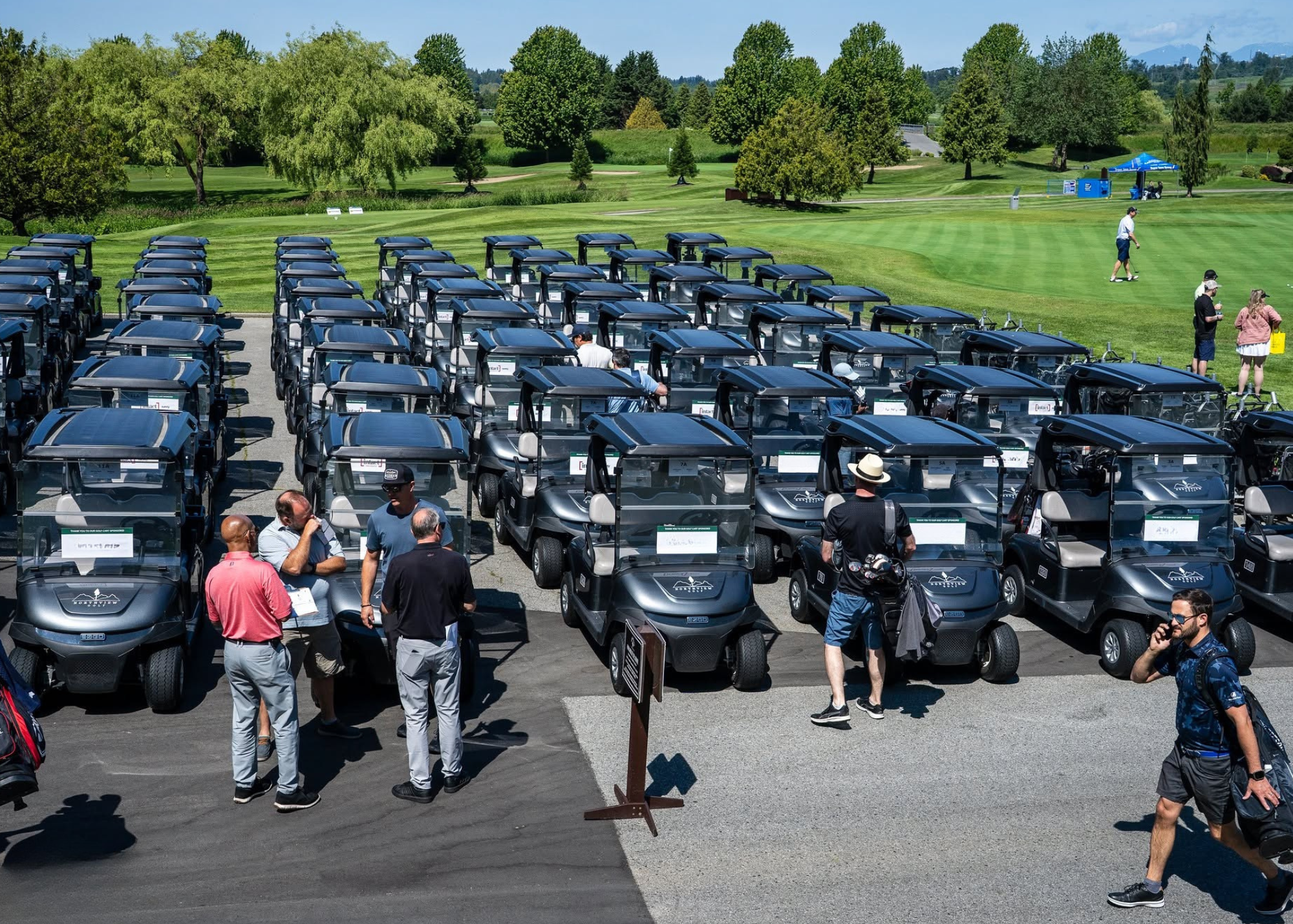 A parking lot filled with black golf carts, with people walking and standing nearby. A golf course with green grass, trees, and a few people playing or walking is in the background.
