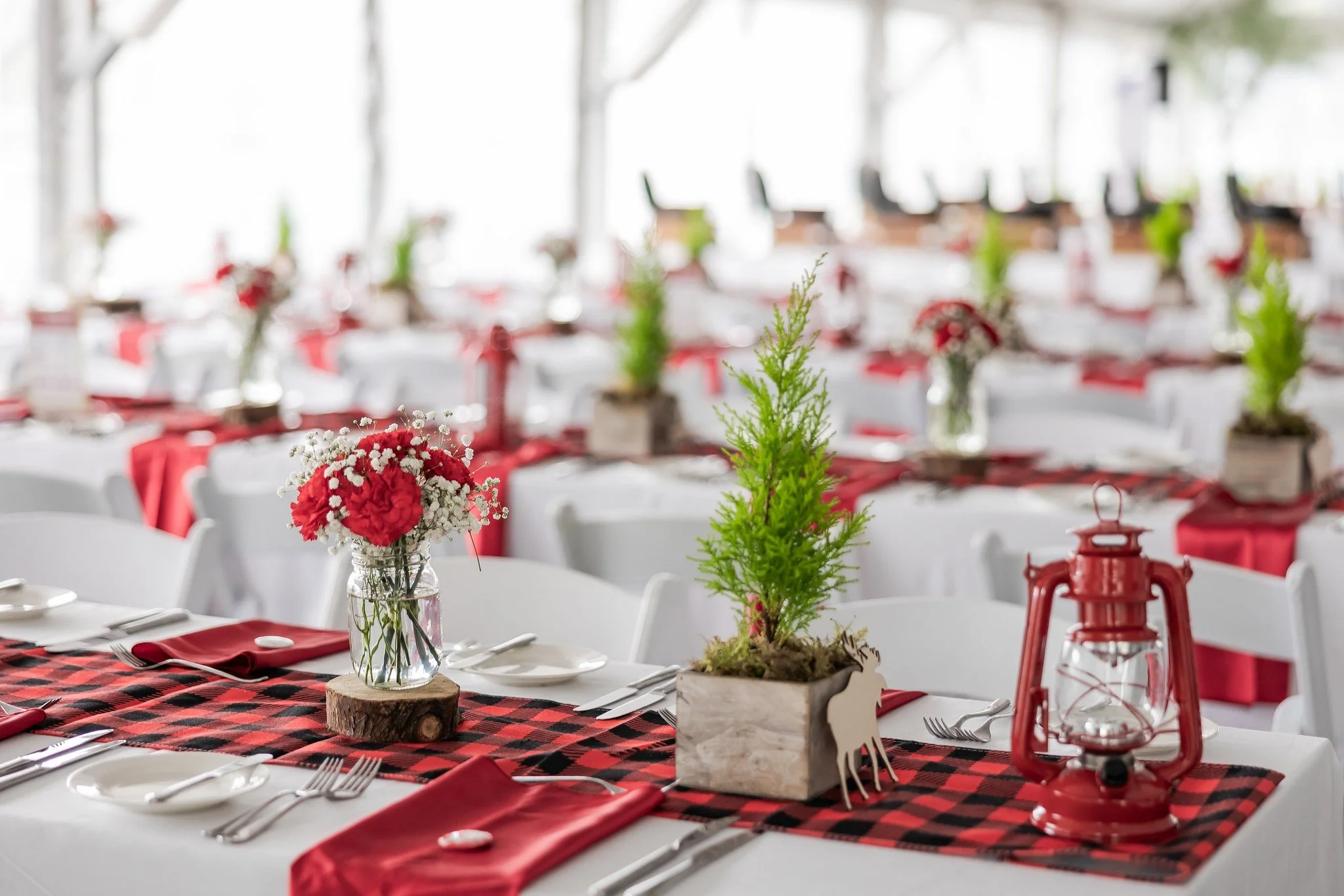 Table setting for a festive event with red and black plaid table runner, white dishes, silverware, red napkins, and holiday-themed centerpieces including small potted pine trees and red and white flower arrangements, along with red lanterns.