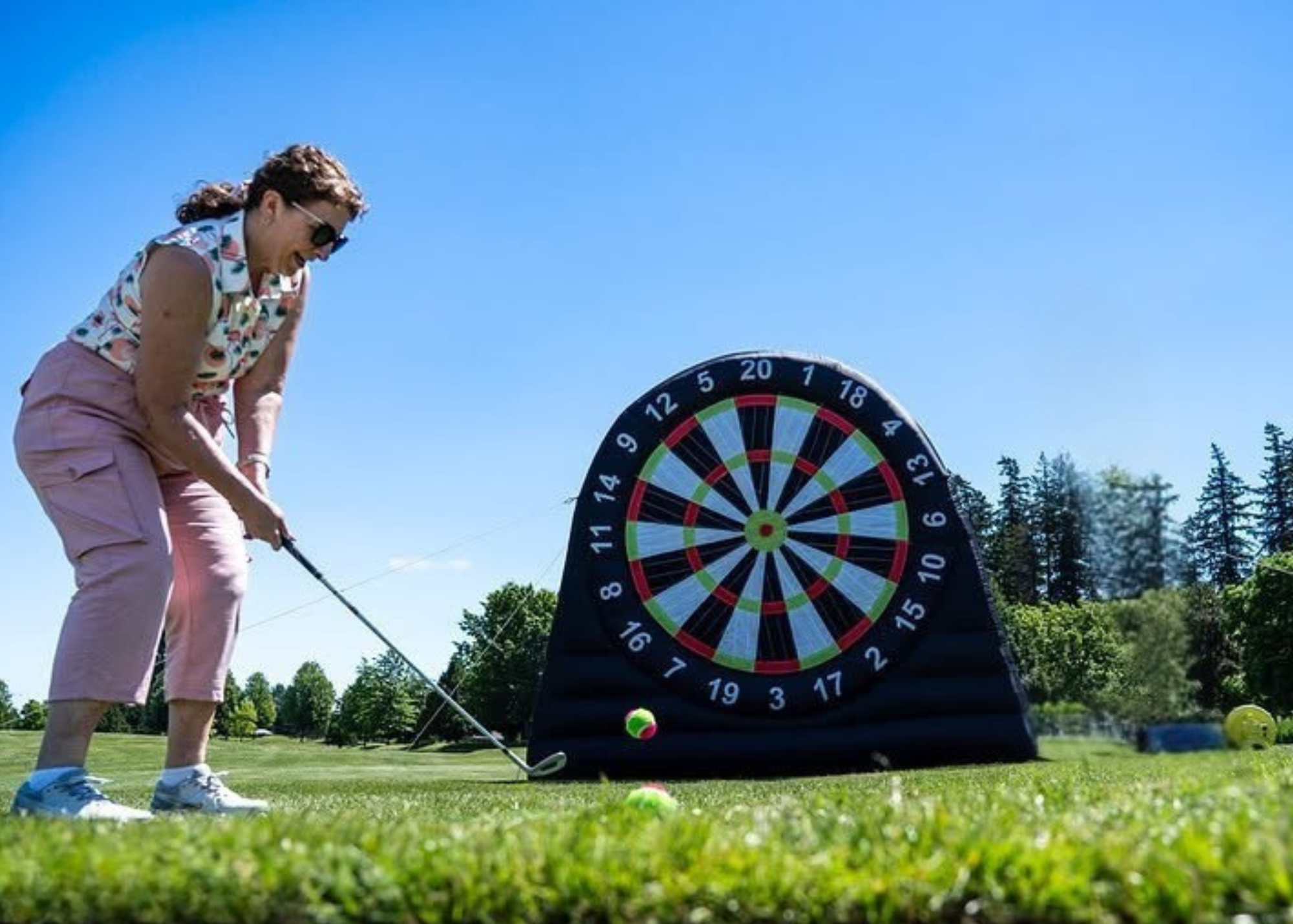 A woman playing golf on a bright, sunny day, wearing sunglasses, a patterned sleeveless shirt, and pink pants, preparing to hit a golf ball on a grassy course next to a large inflatable dartboard.