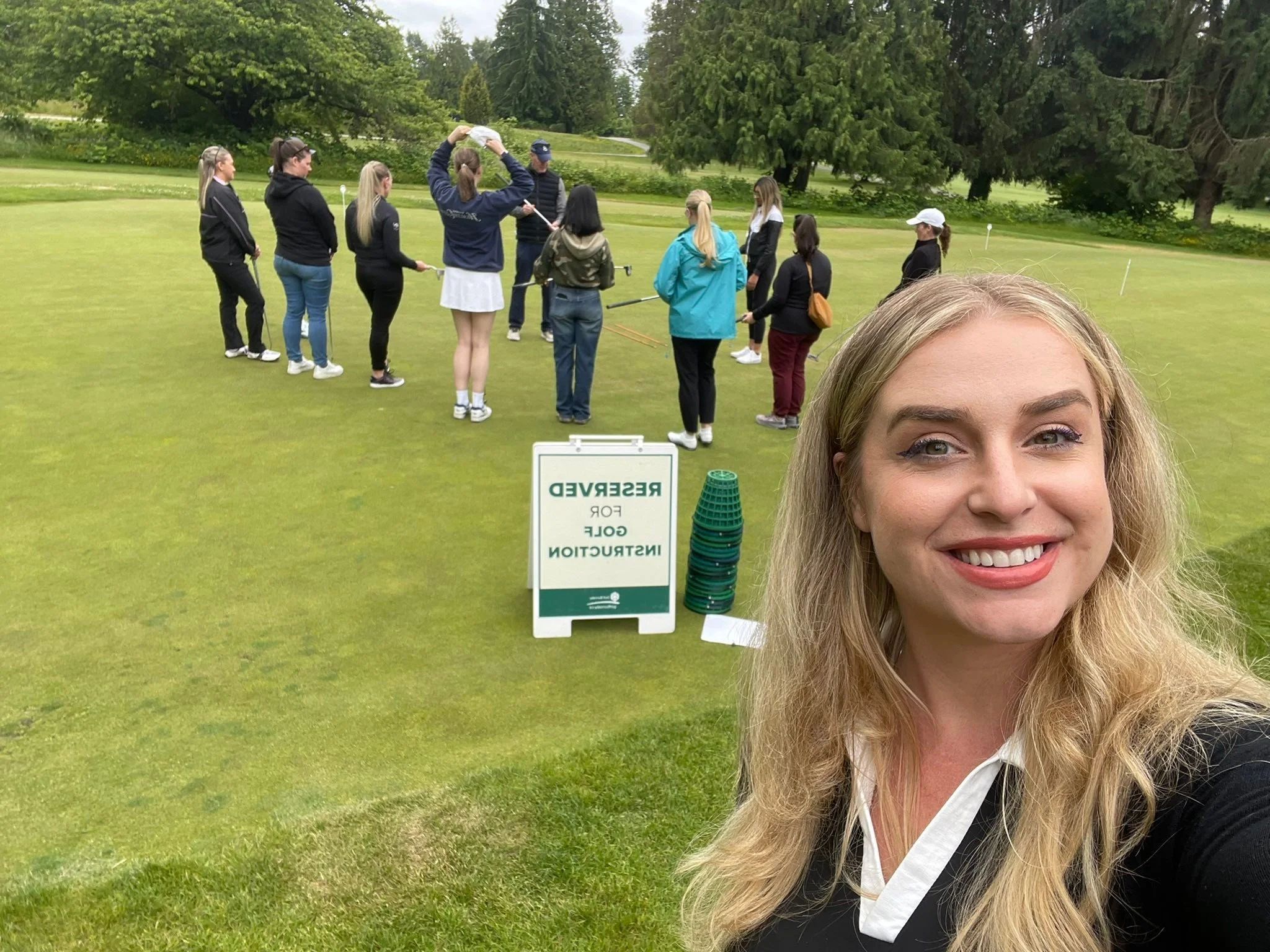 A woman taking a selfie on a golf course with a group of people practicing putting in the background. There is a sign that reads 'RESERVED FOR GOLF INSTRUCTION' and a stack of green cups next to it.