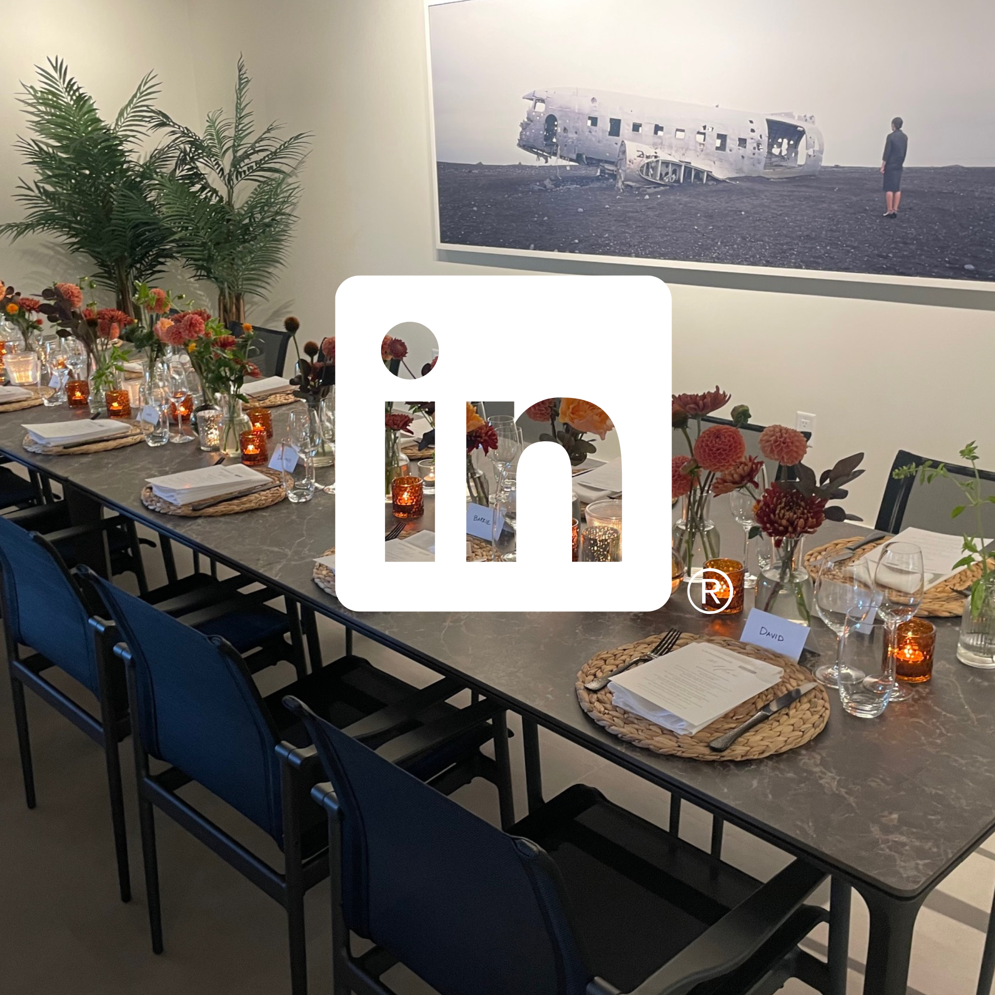 A decorated dining table with pink and orange flower centerpieces, candles, and place settings on woven placemats. A large painting of a plane wreck is on the wall in the background.