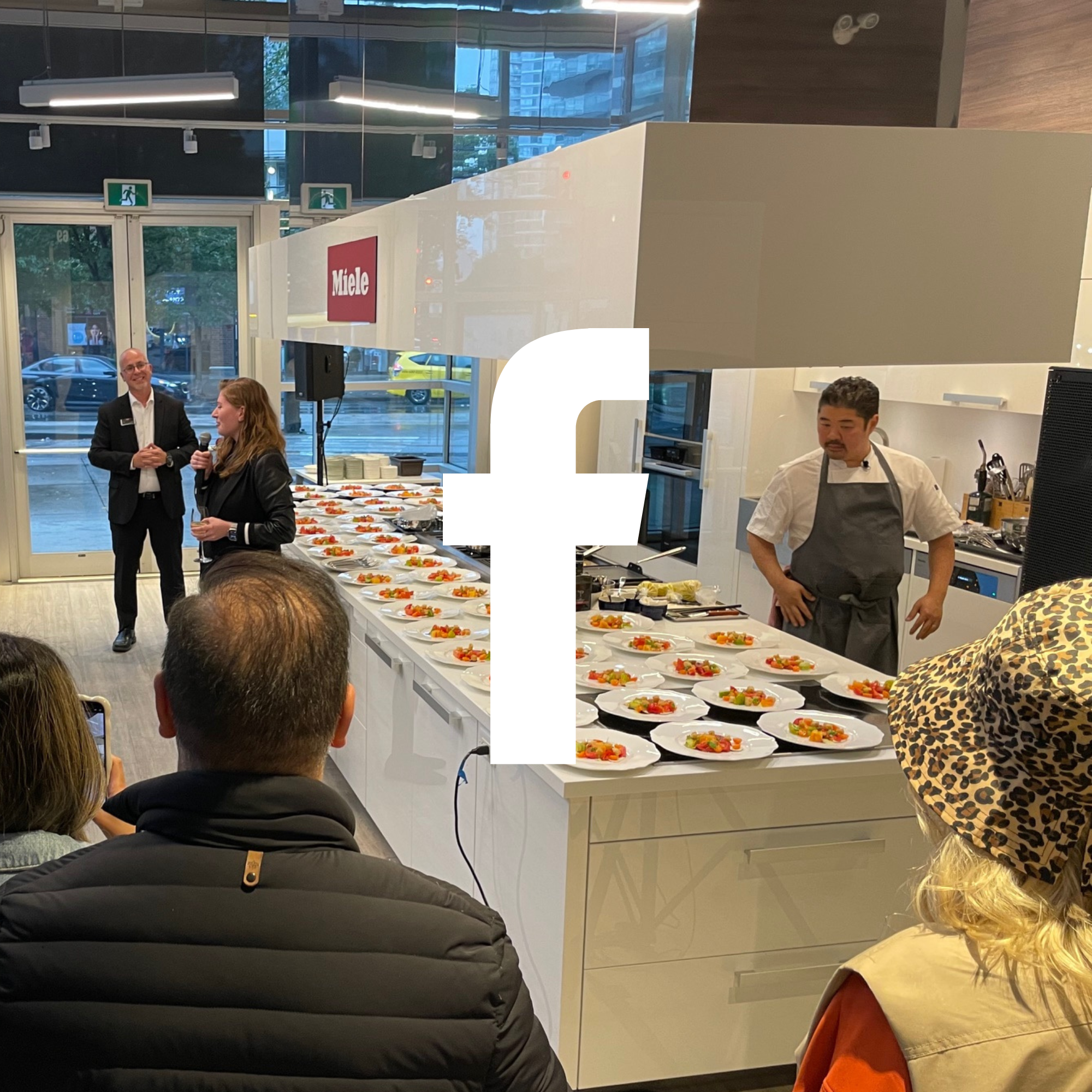 A cooking demonstration or food presentation event in a modern indoor space with a chef, audience, and a table of prepared dishes. There are people speaking at the front, with plates of food arranged on a white counter and a large Facebook logo overlay in the center of the image.
