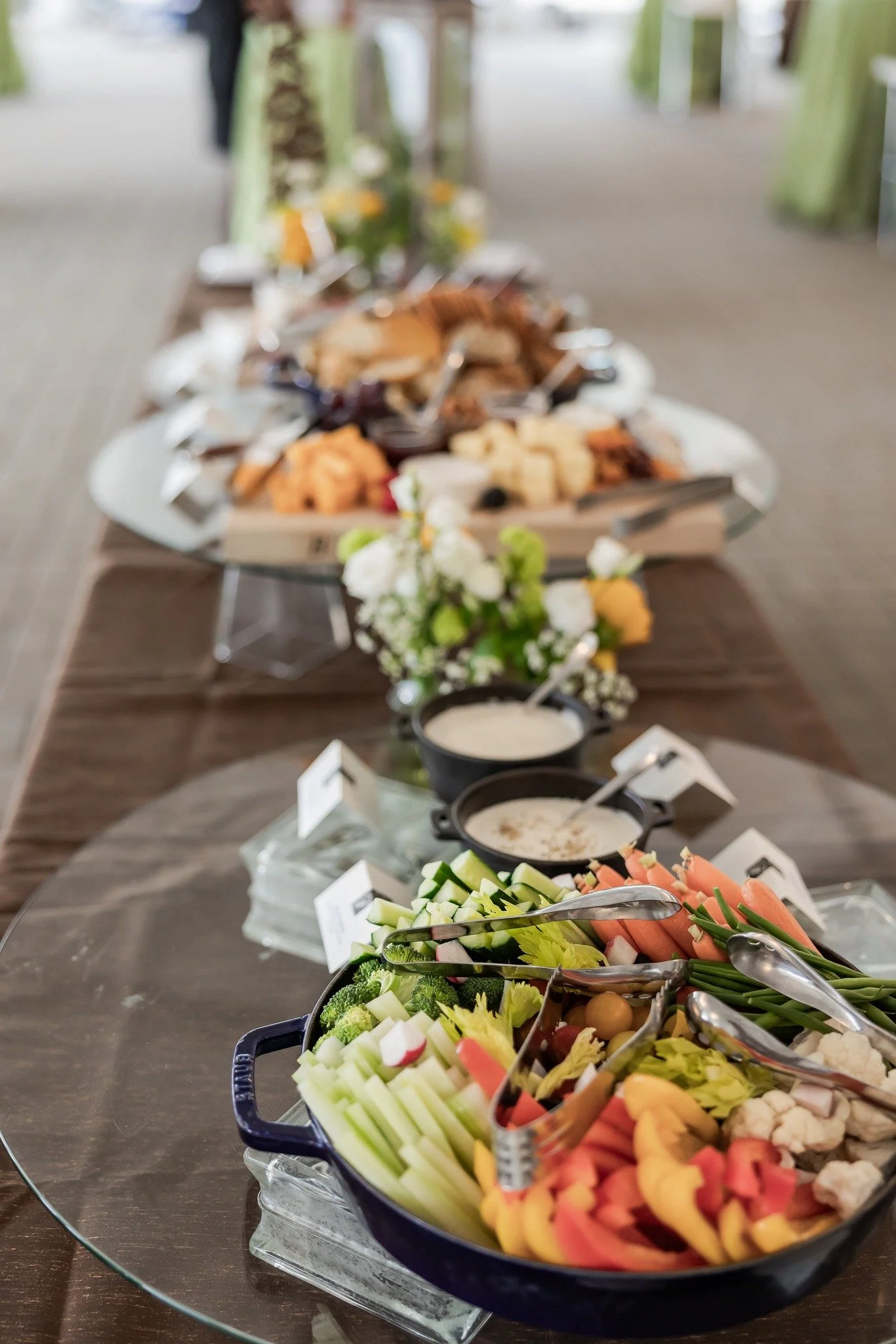 Vegetable platter with cucumber, carrots, broccoli, cauliflower, and cherry tomatoes, along with sauces in small bowls, set on a glass tray at a buffet table.