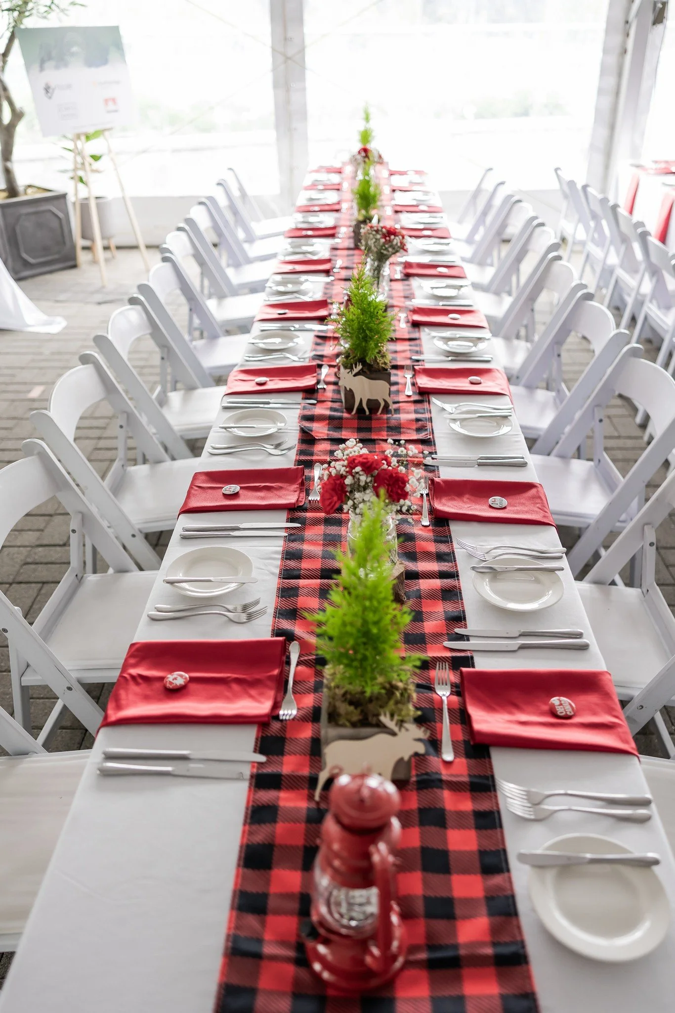 Long banquet table decorated for a holiday celebration with red and black plaid table runner, white plates, silverware, red napkins, and small green Christmas trees in pots along the center, accented with red flowers in vases and decorative reindeer.