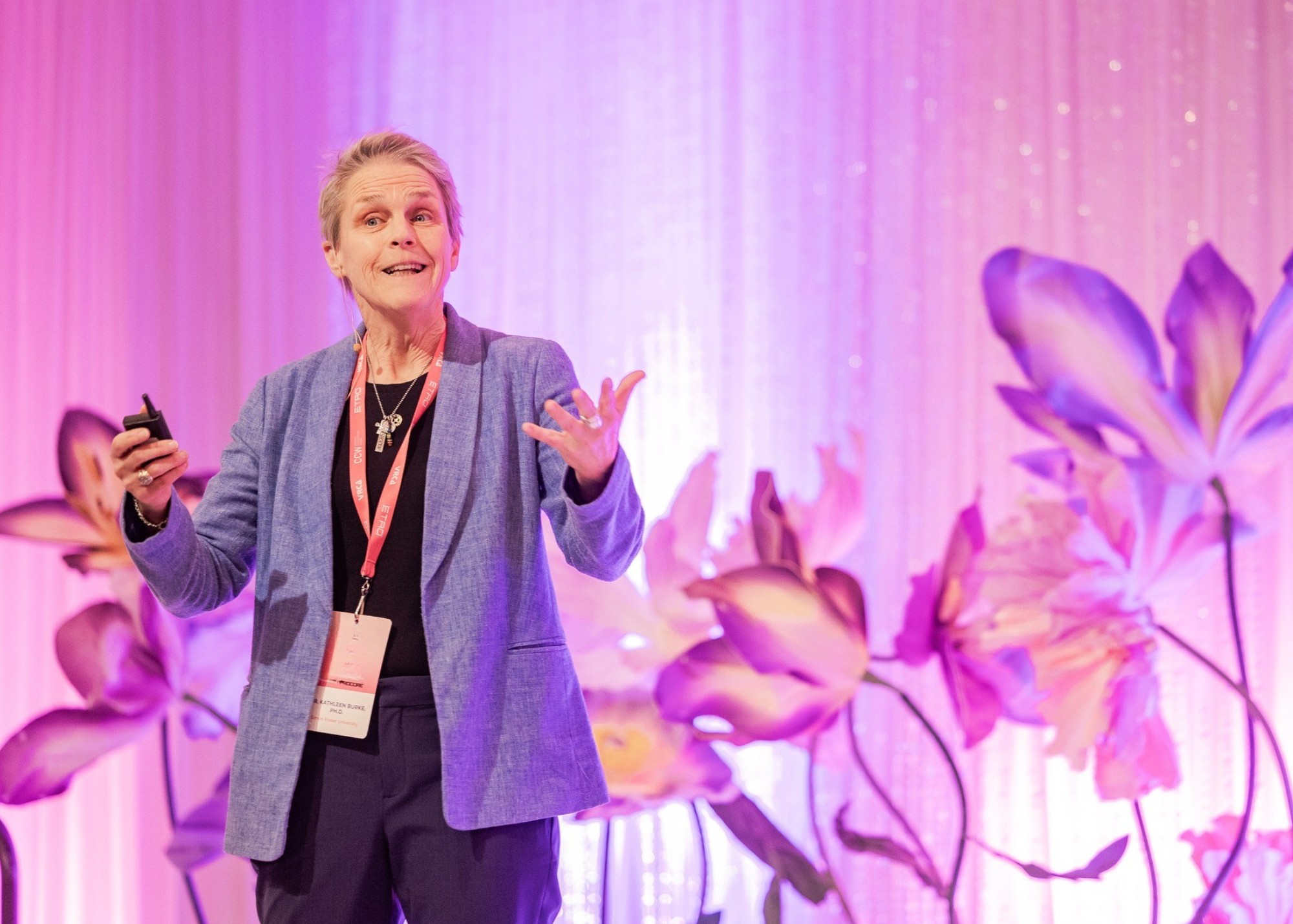 A woman giving a presentation on stage with purple and pink floral decorations in the background.