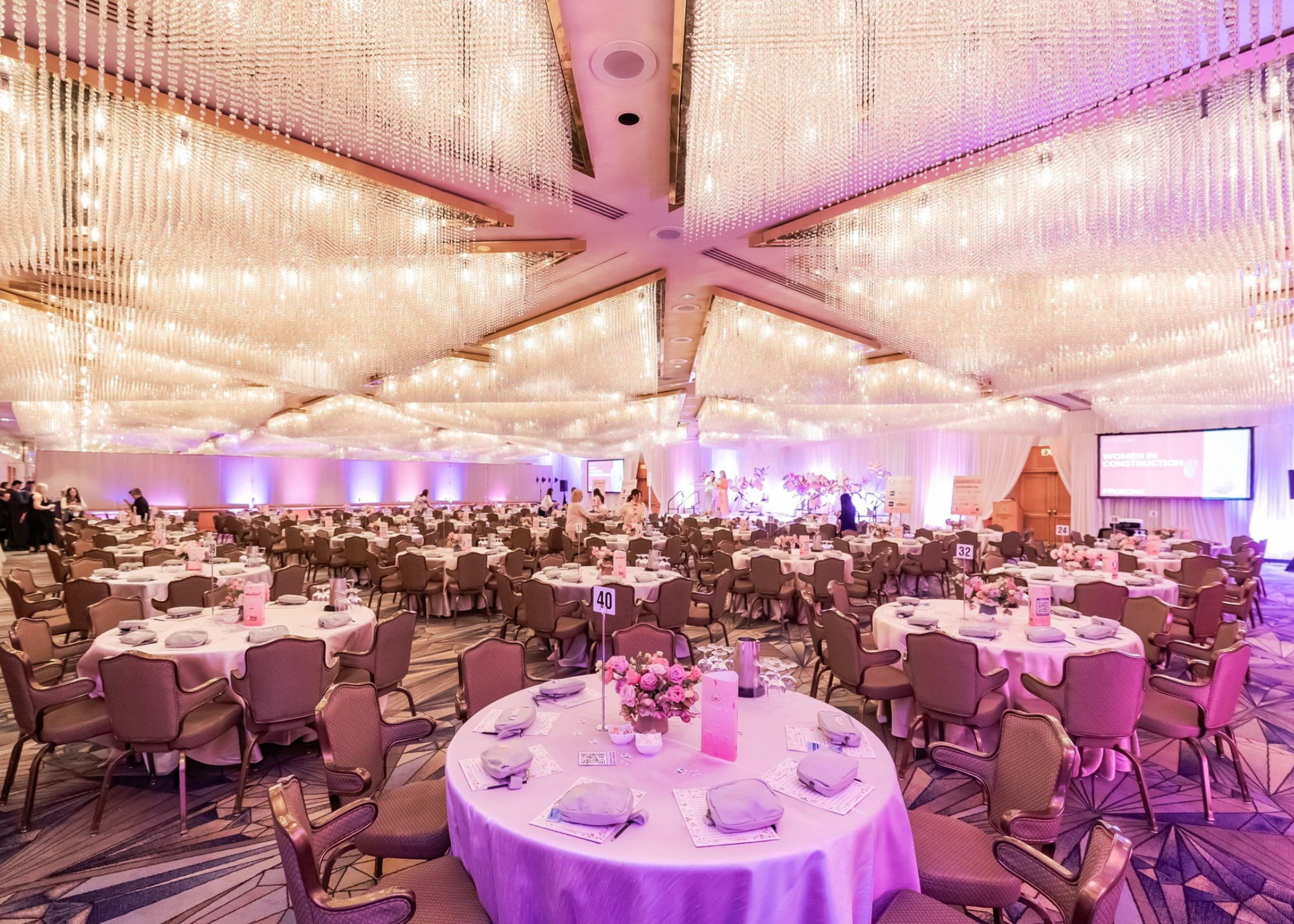 A large banquet hall decorated with pink lighting, filled with round tables set for an event, with centerpieces, napkins, and place settings, and a stage in the background with people preparing for a presentation or performance.