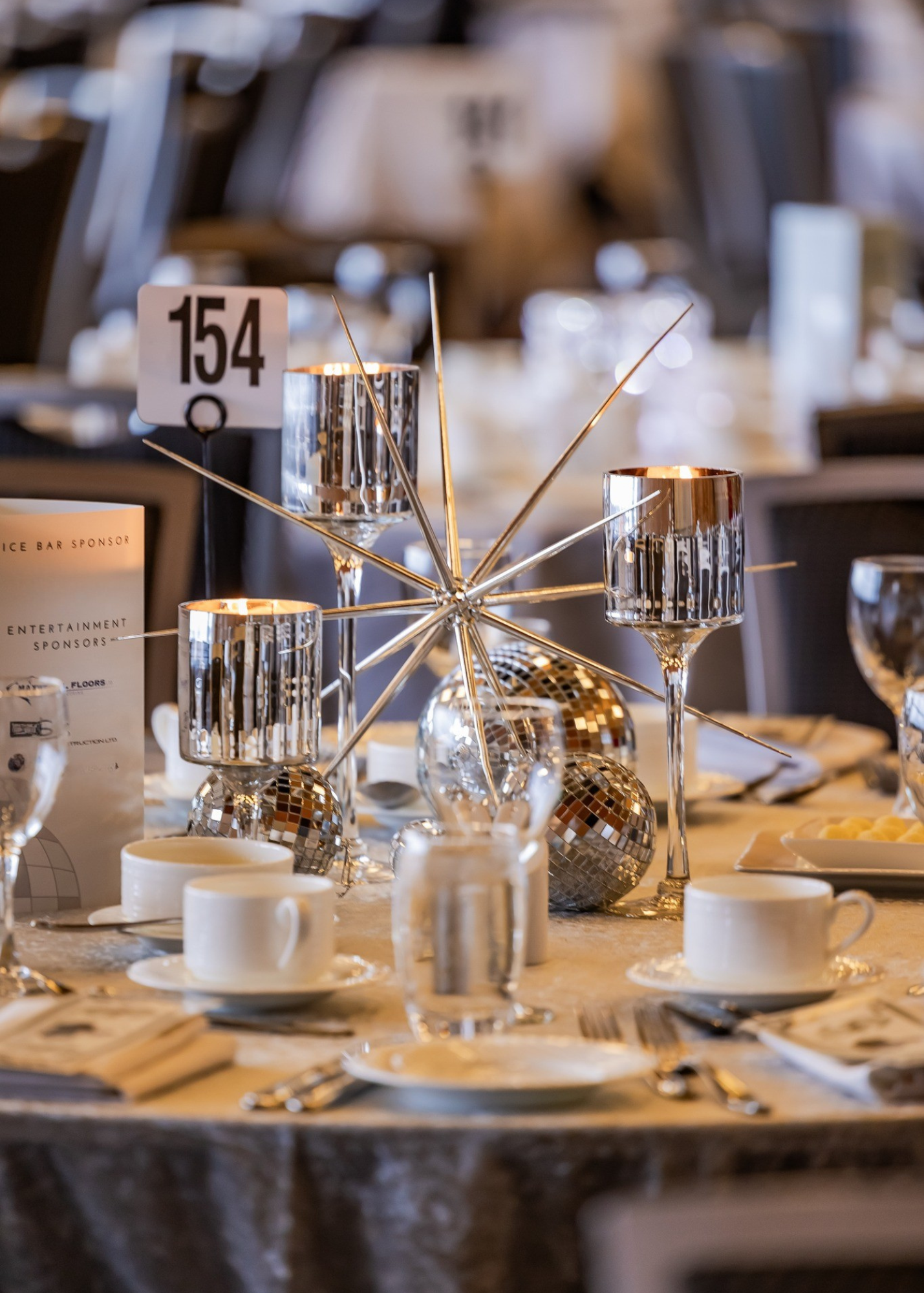 A table decorated with silver and gold geometric ornaments, including a starburst centerpiece and disco ball accents, set for a formal event with white teacups, plates, silverware, and glasses.