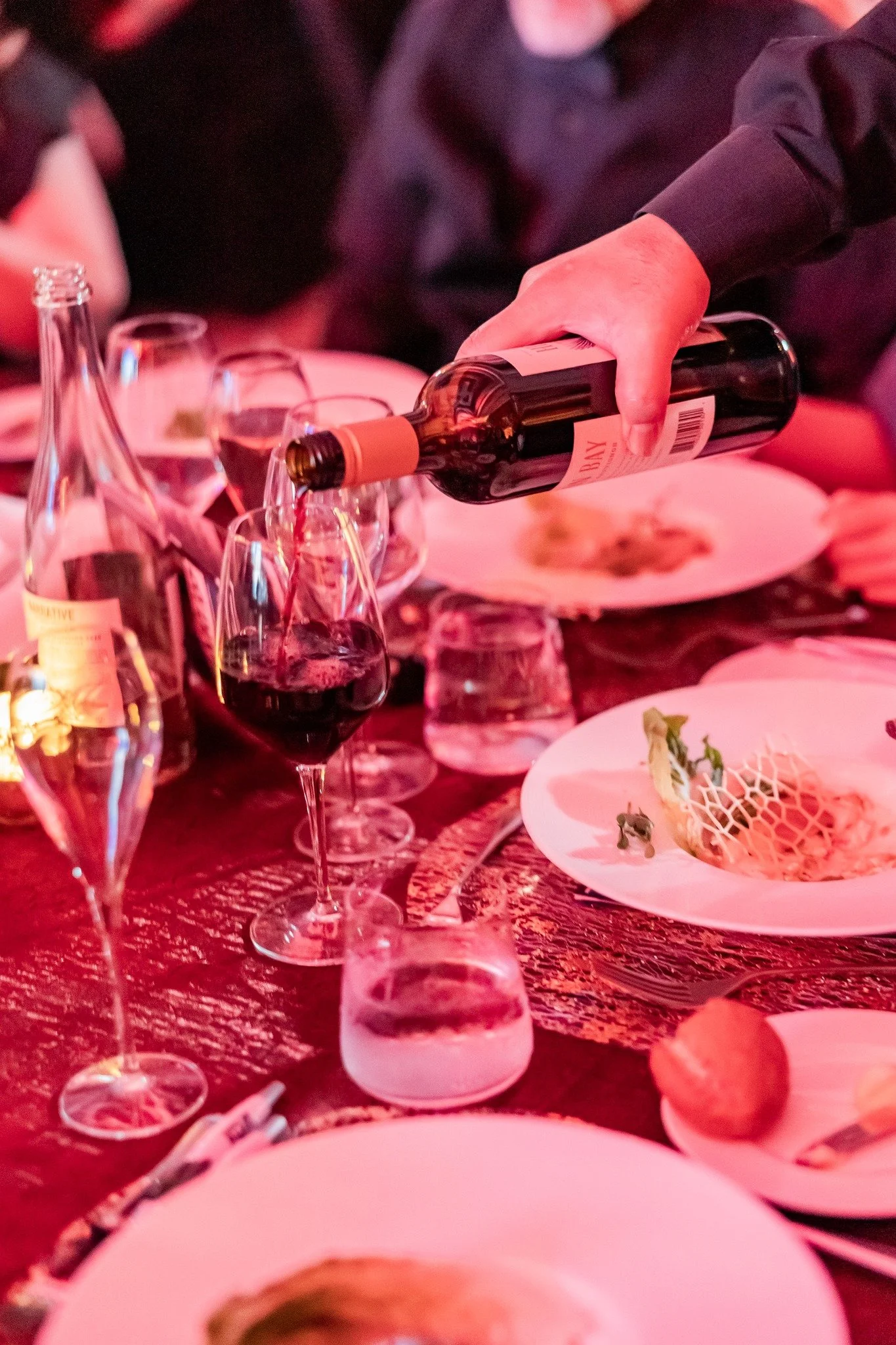 Person pouring red wine into a glass at a dinner table with plates, glasses, and food.
