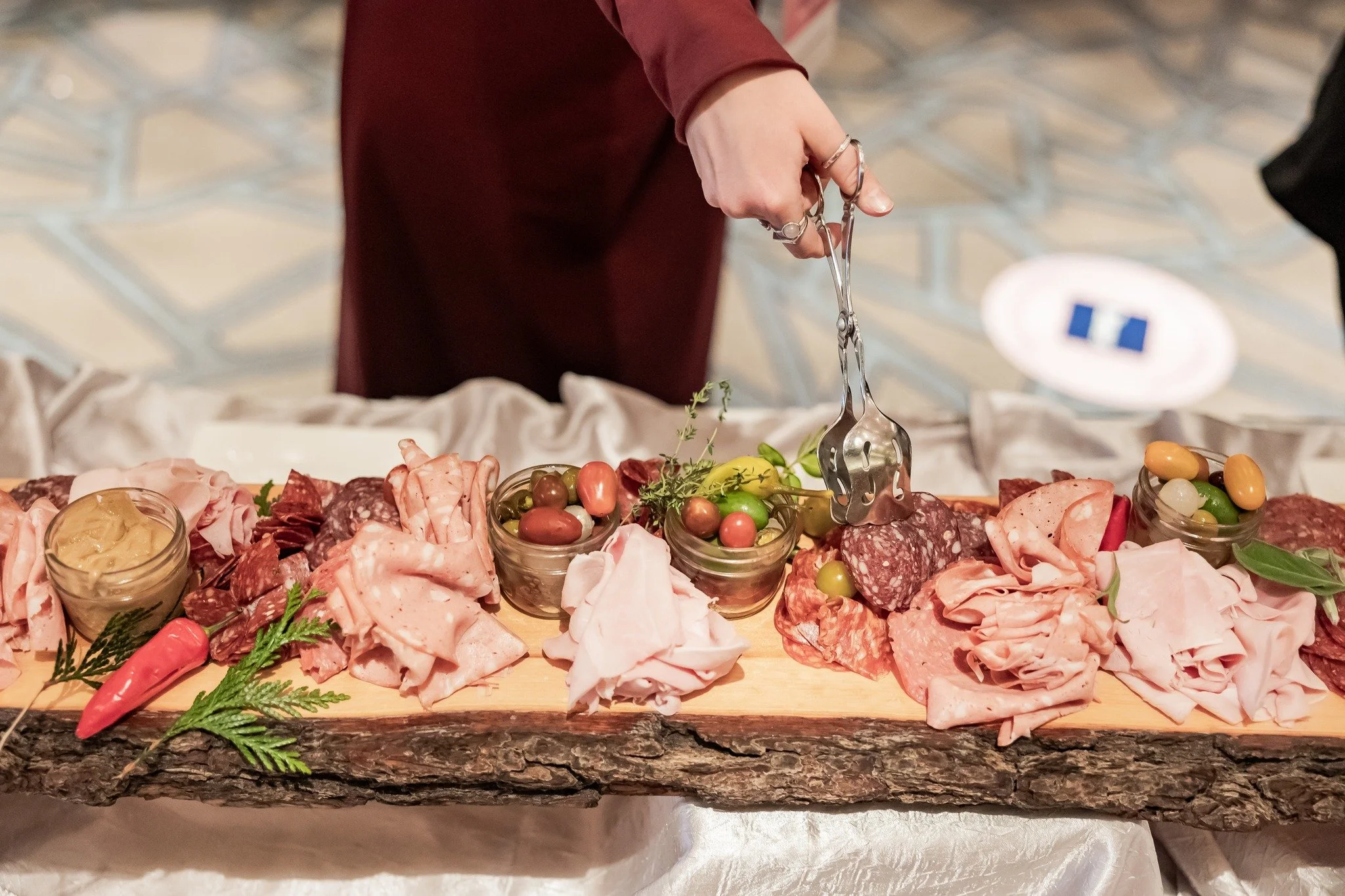 A person slicing cured meats on a rustic wooden platter with olives, pickles, and garnishes.