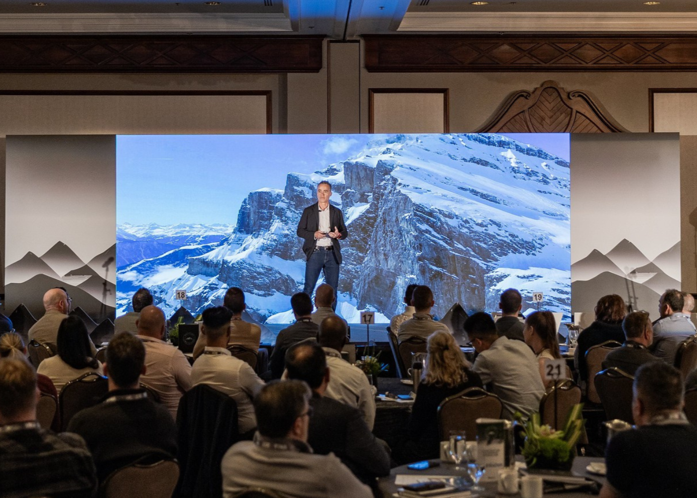 A man in a dark blazer and white shirt giving a presentation on stage at a conference, with a large mountain landscape screen behind him, showing snowy peaks and cliffs, and an audience seated at round tables.