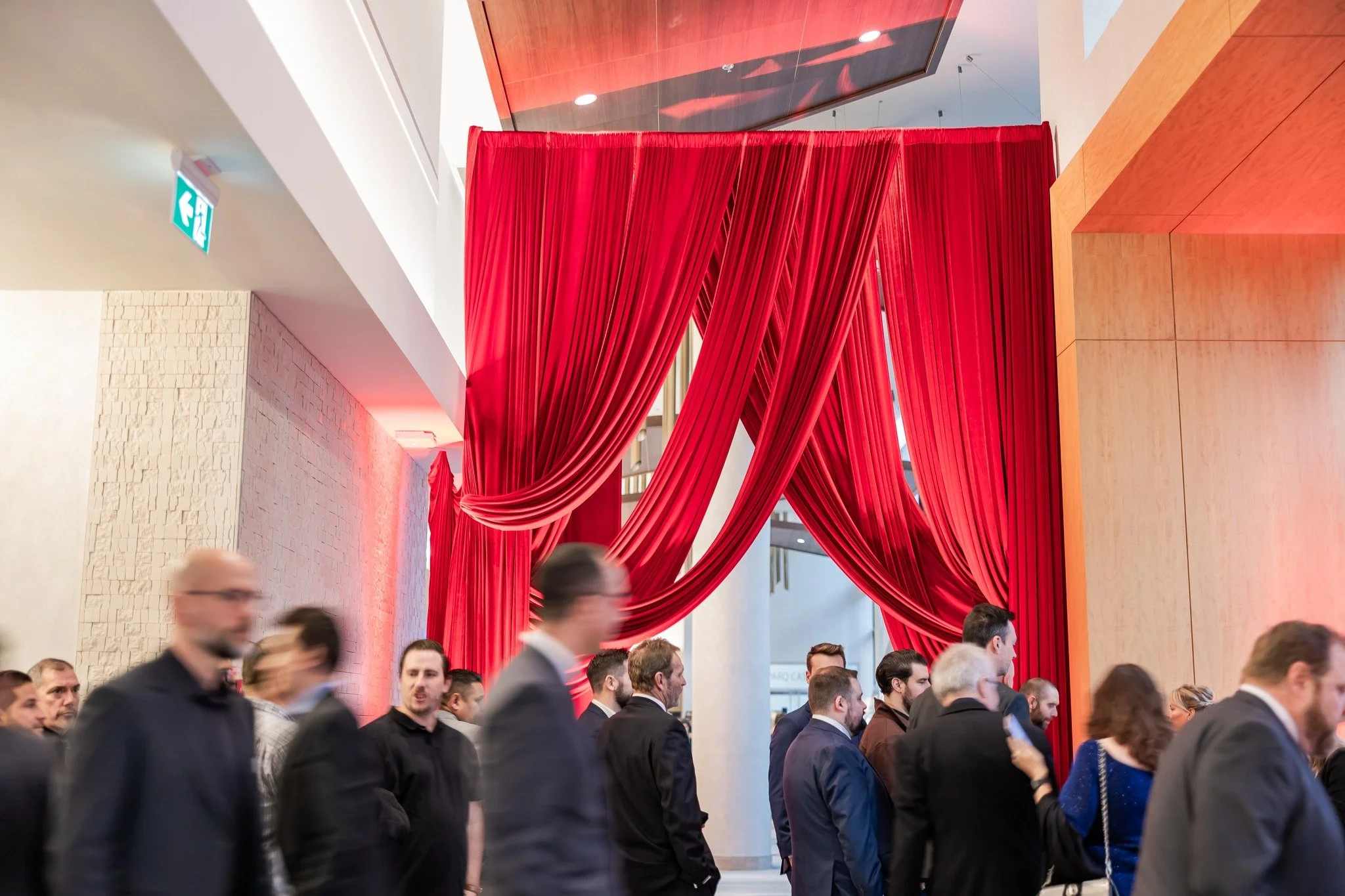 People standing in line or waiting at an indoor event, with large red curtains hanging in the background.