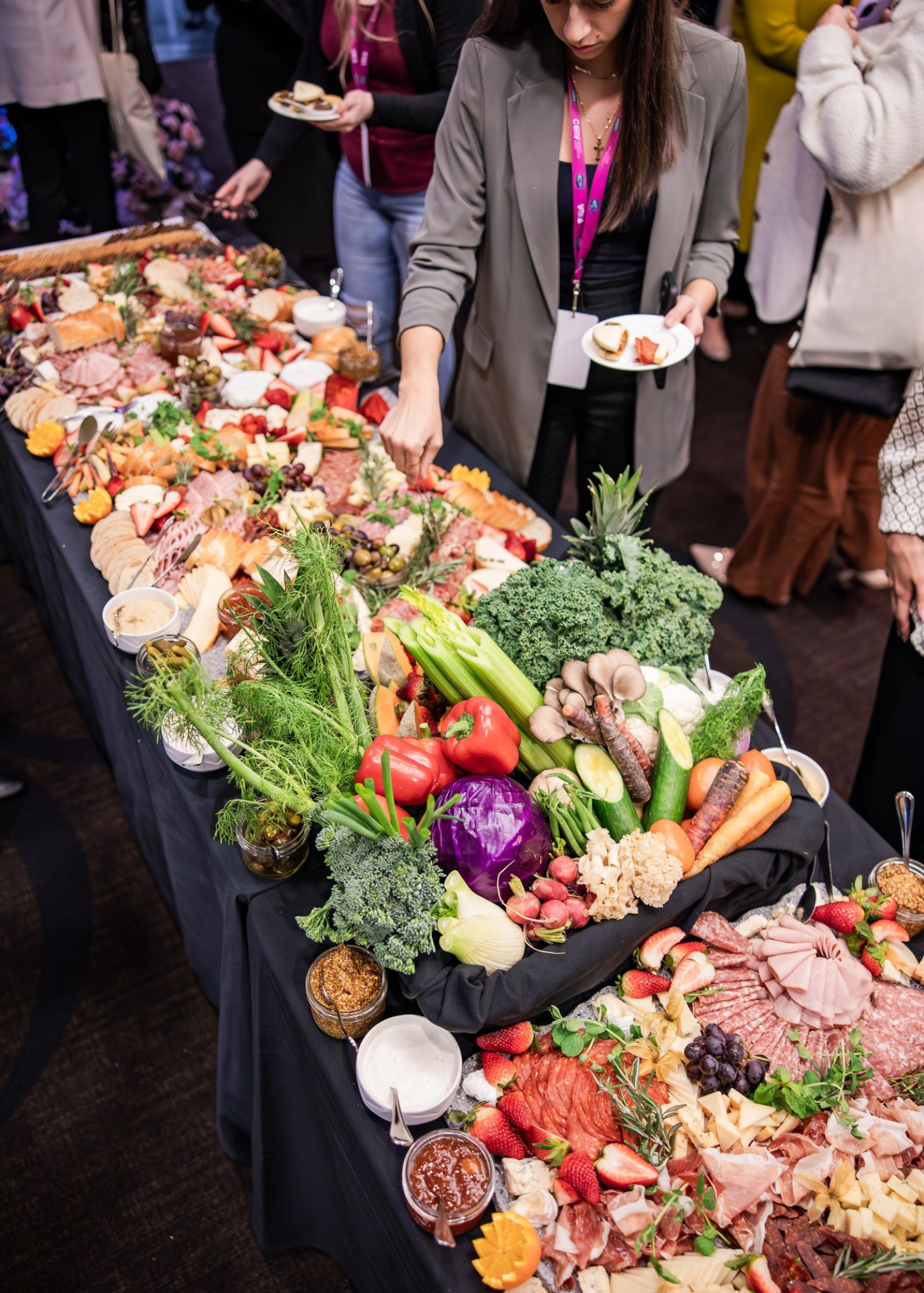 A buffet table filled with fresh vegetables, fruits, and cold cuts at a social gathering. Several people are serving themselves or holding plates.