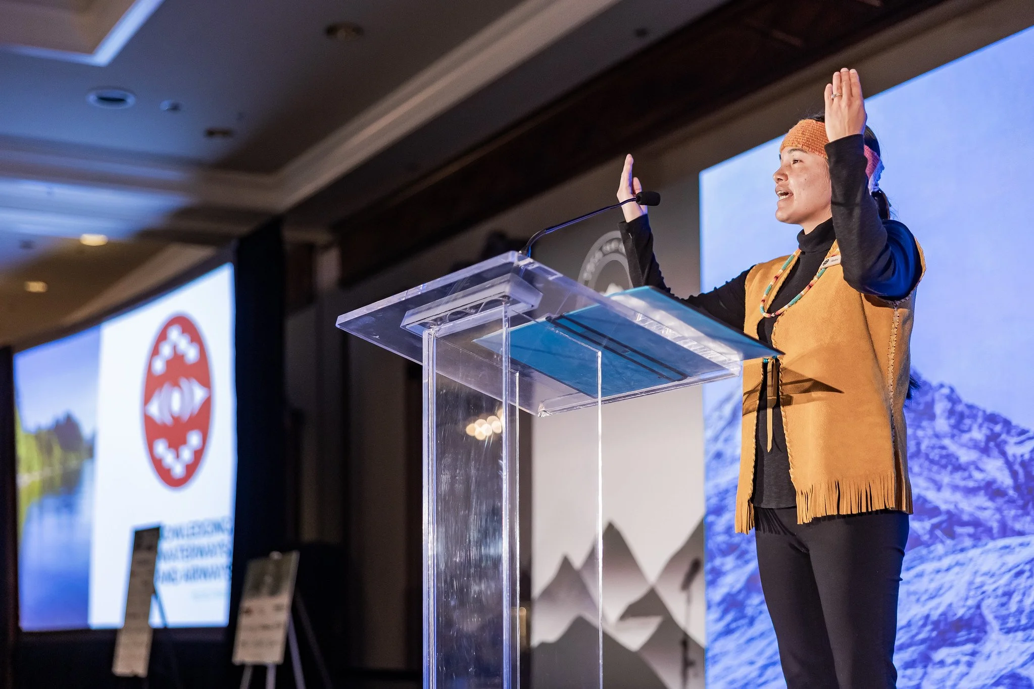 A Indigenous Woman speaking  and providing a cultural welcome at a podium during a conference or event, with large screens displaying mountain scenery and a logo in the background.