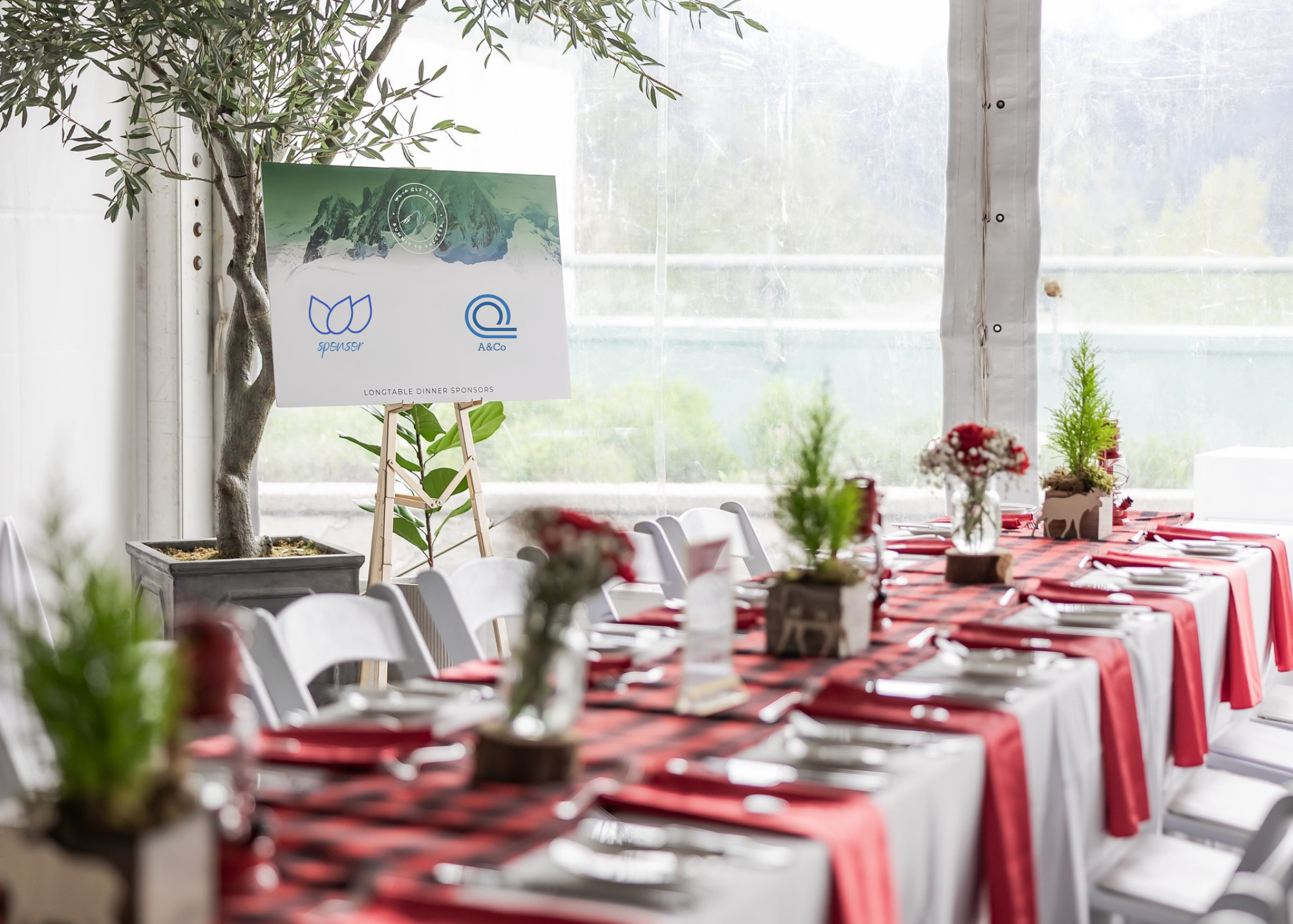 Interior of a decorated banquet tent with a long table, white chairs, red and black checkered tablecloths, flower centerpieces, and a forest-themed sponsor display on an easel