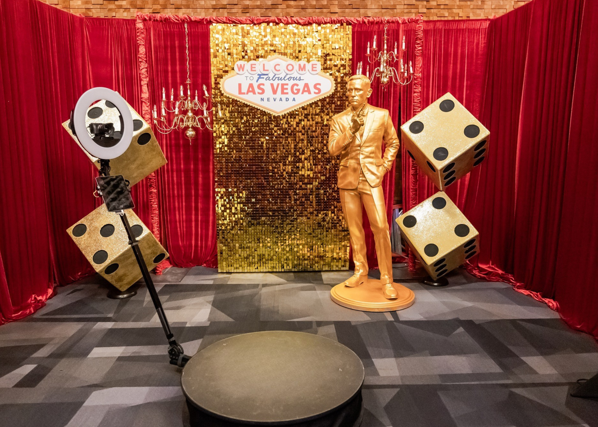 A Las Vegas themed photo setup with a golden statue of a man, red curtains, and large dice decorations. Also includes a sign that says "Welcome to Fabulous Las Vegas Nevada" and a ring light with a smartphone attached for photography.