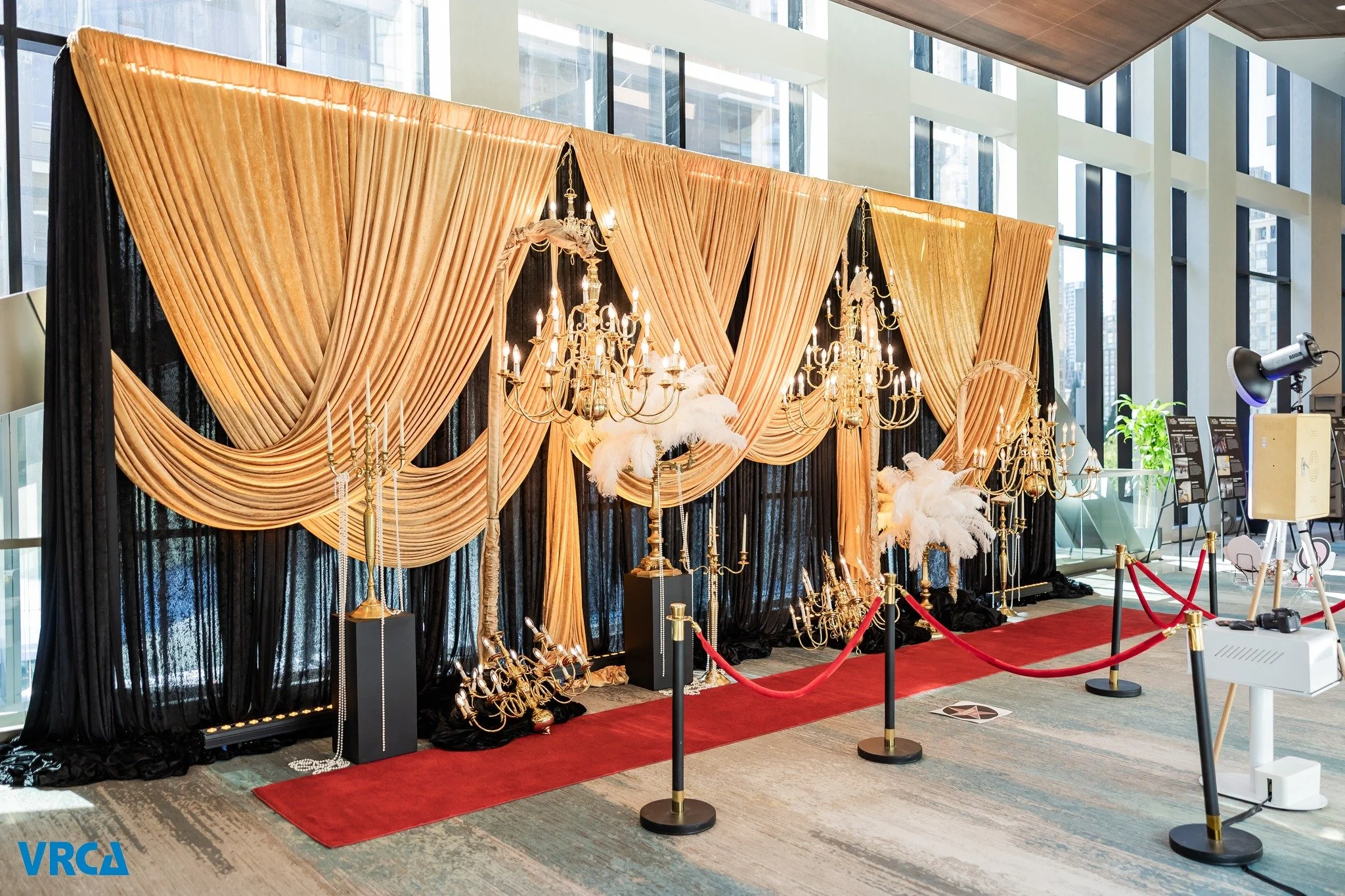A decorative backdrop for photos with orange and black curtains, ornate chandeliers, and candelabras, set up for an event or photo booth, with a red carpet, ropes, and a photo booth camera on a tripod in a modern building interior with large windows.