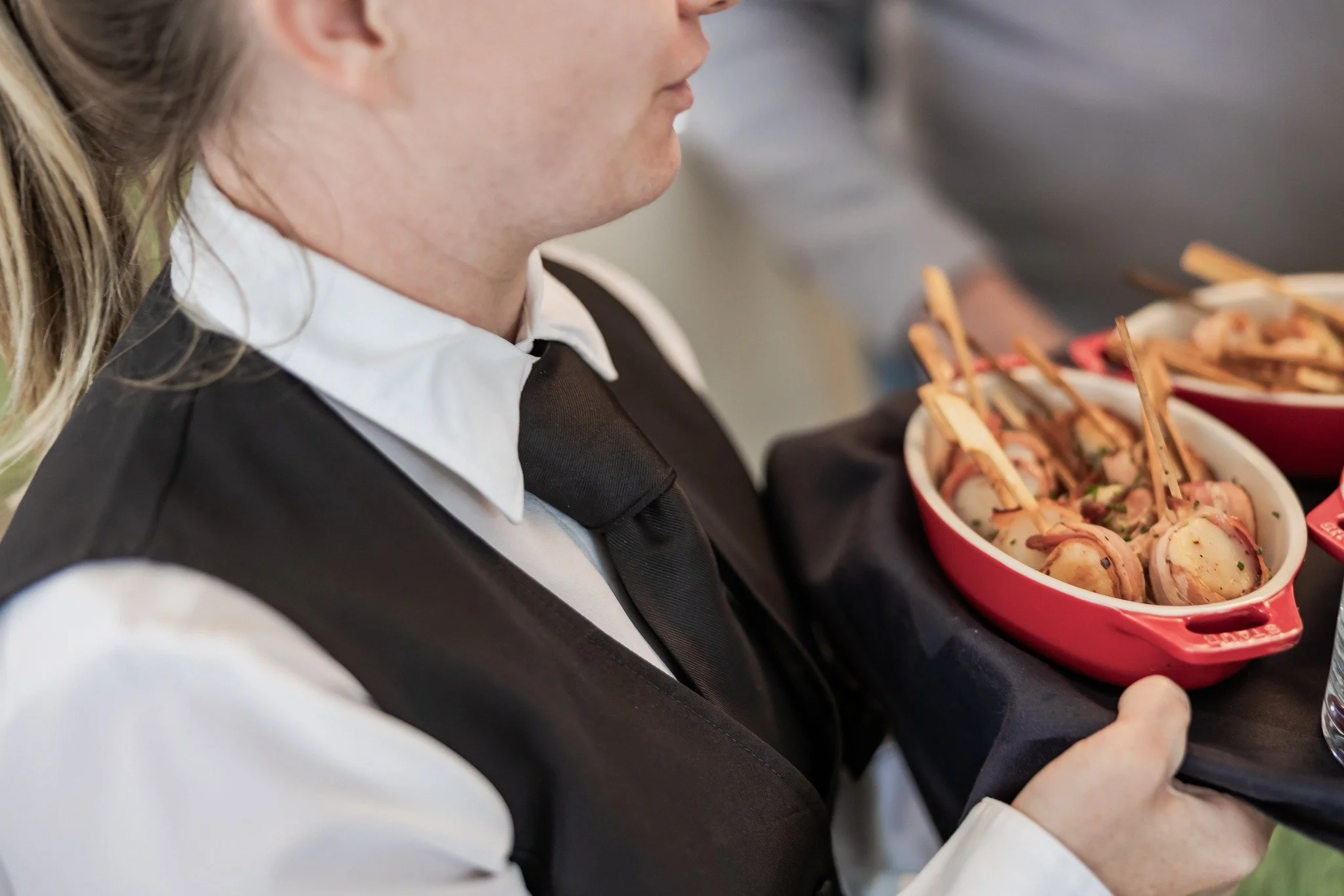 A waitress in a black vest and white shirt holding a red bowl of sliced open-faced sandwiches decorated with herbs.