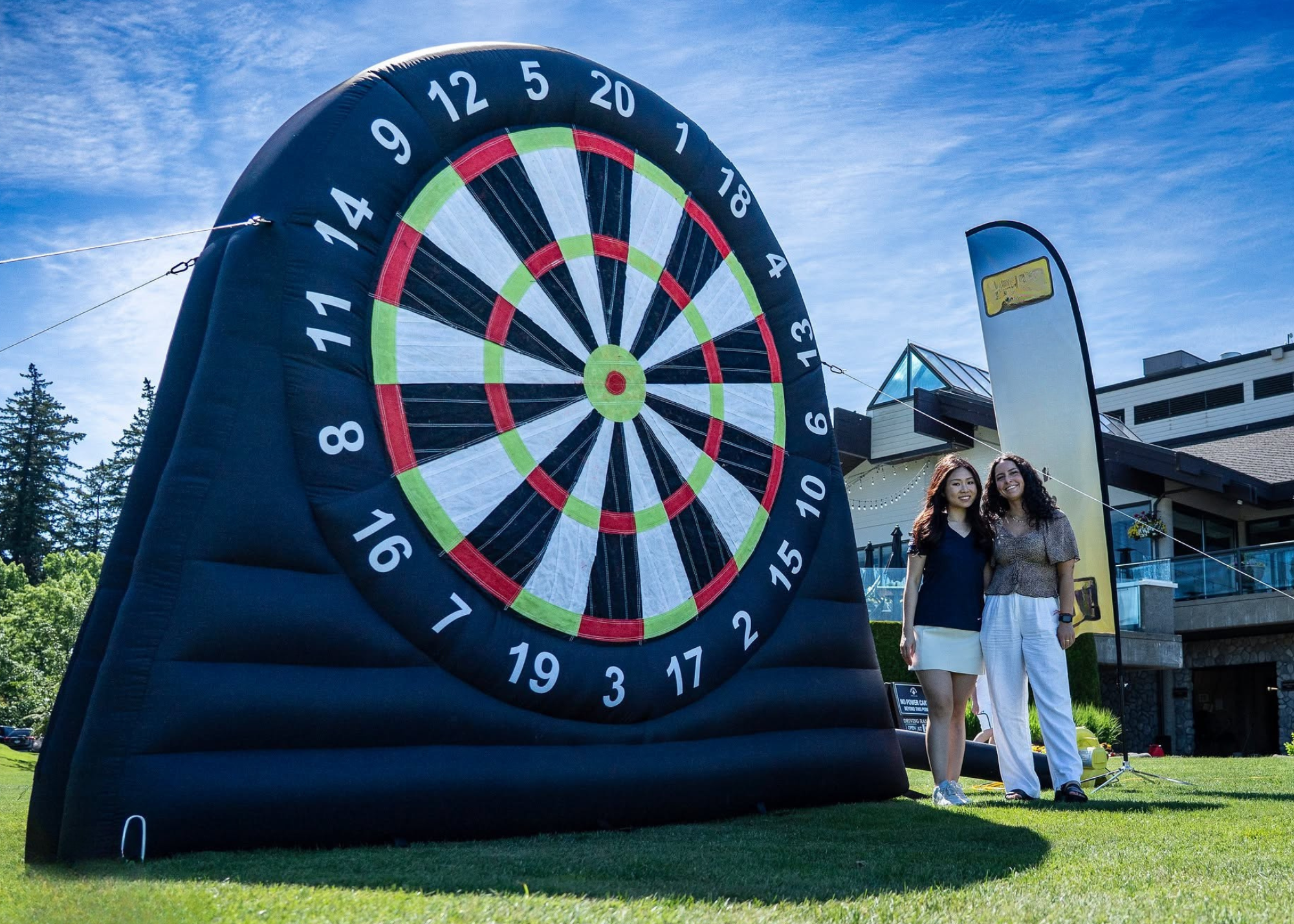 Two women standing next to a large inflatable dartboard outdoor on a grassy area with a building in the background and clear blue sky.