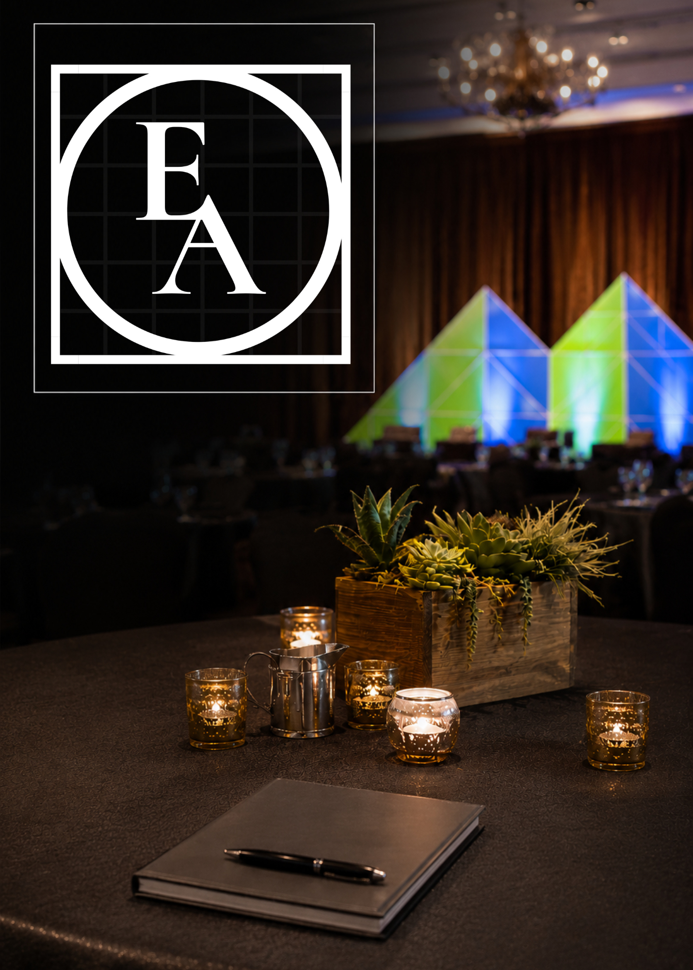 Decorative conference or event setup with table, candles, succulent centerpiece, and a large illuminated geometric art installation in the background, with a sign or logo featuring the letters 'E' and 'A' enclosed in a circle and square.