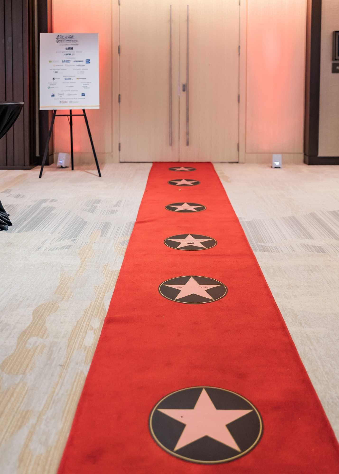 Red carpet with Hollywood star footprints leading to closed doors, with a poster on an easel and soft lighting in the background.