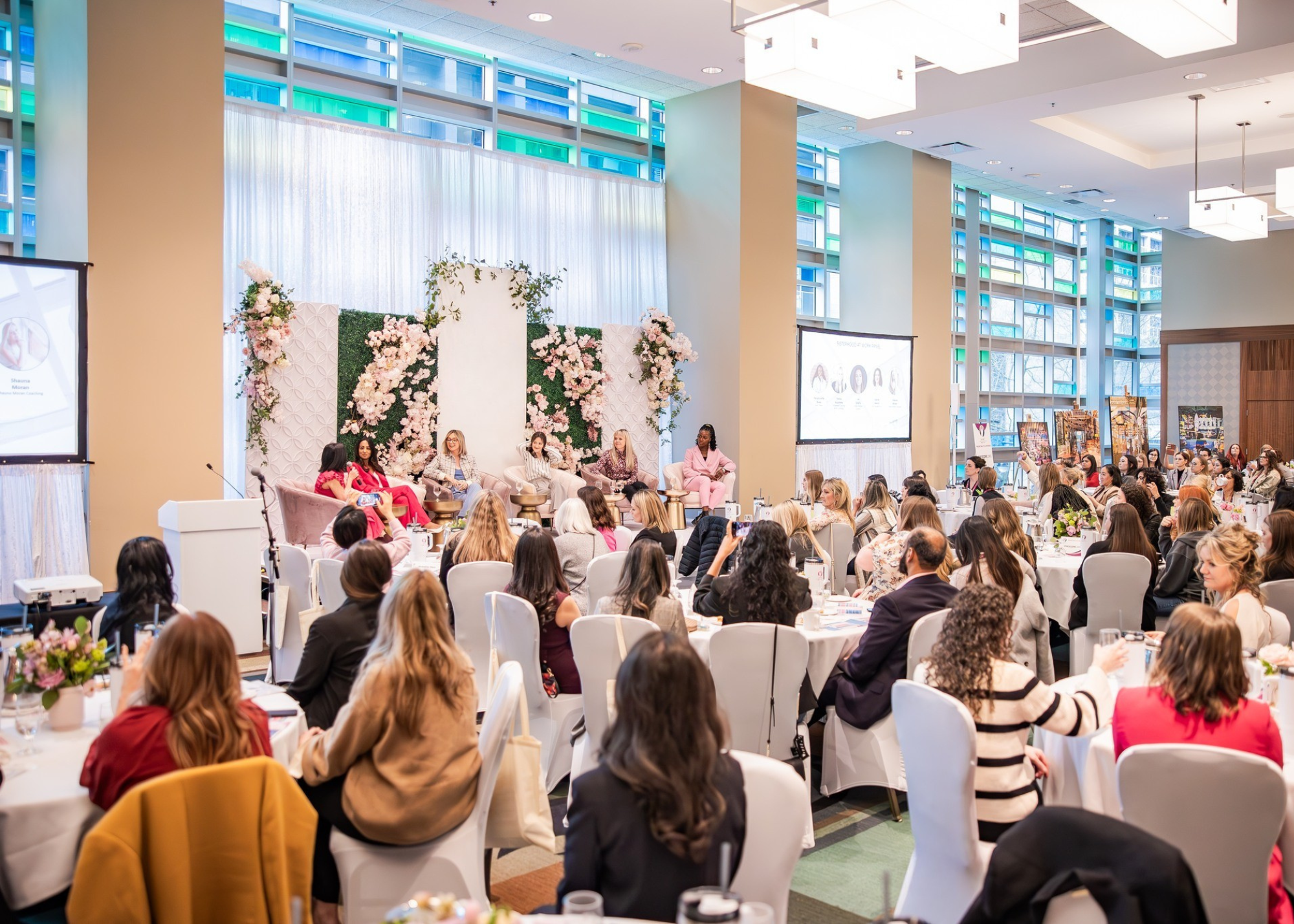 A conference room filled with women seated at tables, listening to a panel of five women on stage with floral decorations behind them, and large screens displaying presentation slides.
