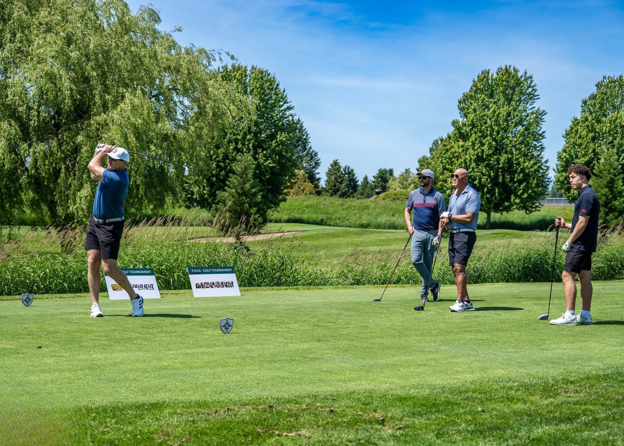 A man playing golf on a sunny day while three others watch on a golf course with trees and grass.