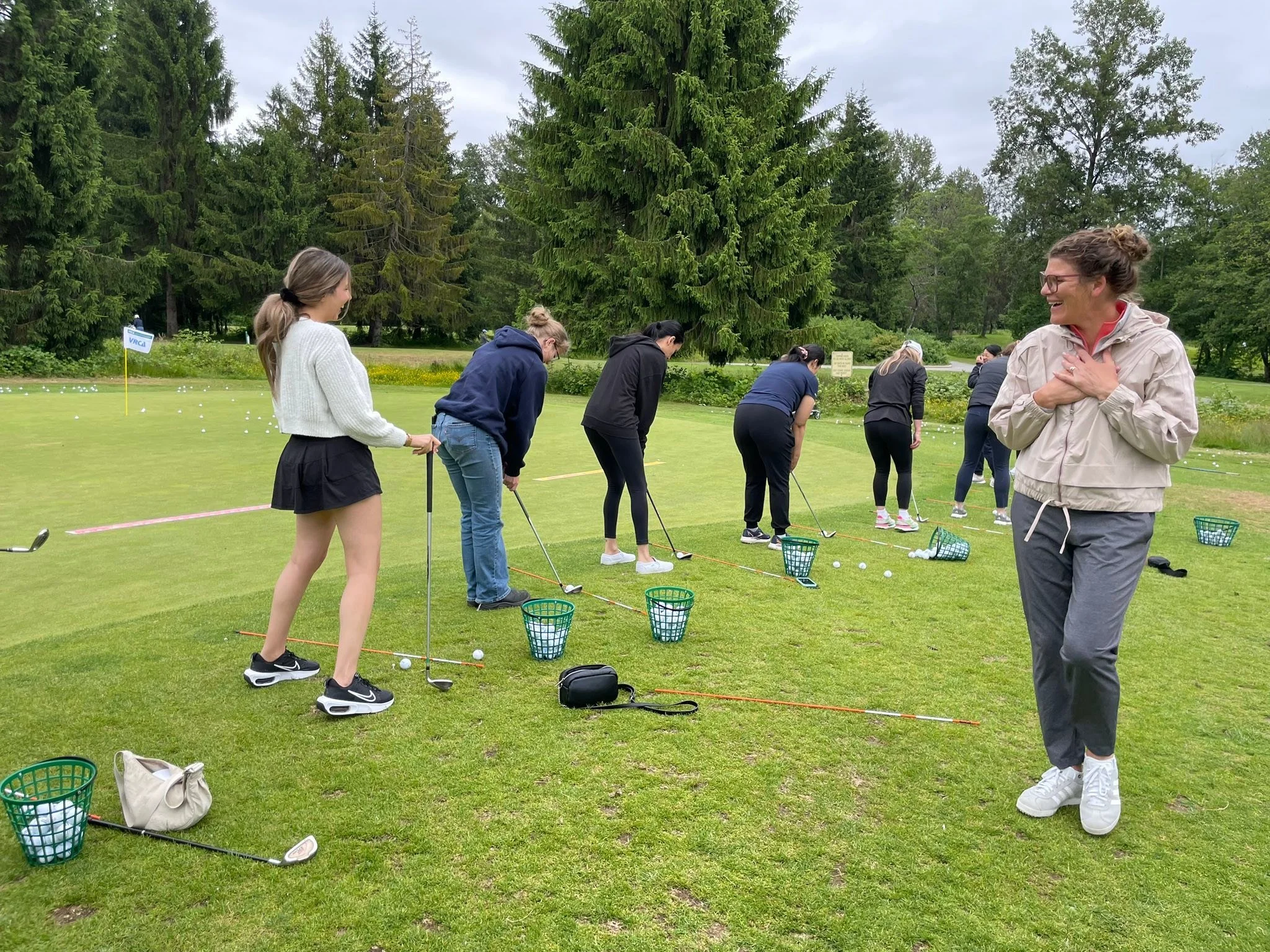 Women practicing golf on a practice range with green grass, trees, and golf balls scattered on the ground.