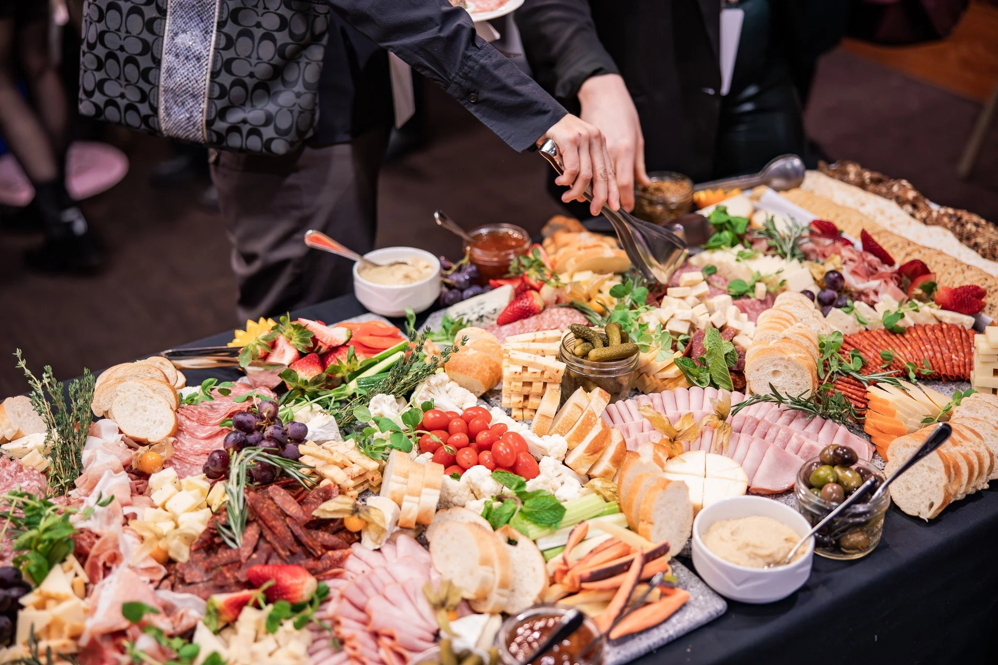 A person is serving themselves from a large charcuterie board filled with assorted cheeses, sliced meats, cherry tomatoes, fresh strawberries, grapes, vegetables, bread, crackers, and various condiments at a social gathering.